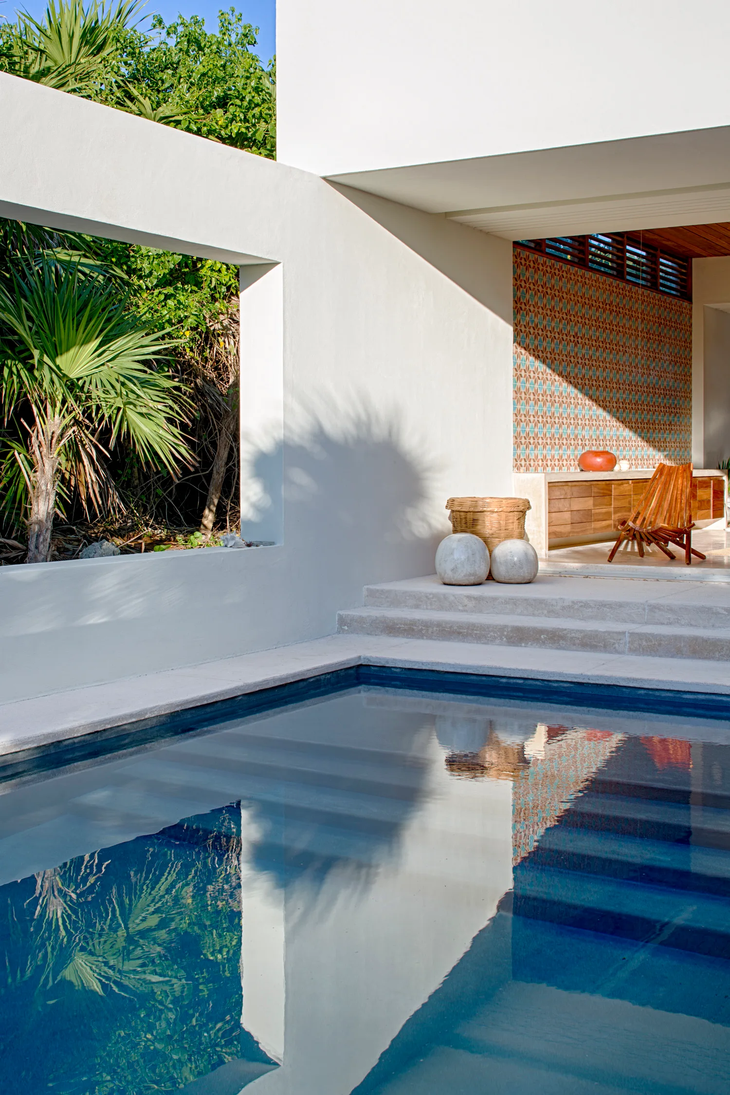 A contemporary swimming pool adjacent to a white concrete patio featuring a wall cutout framing green foliage and a covered area with patterned tile.