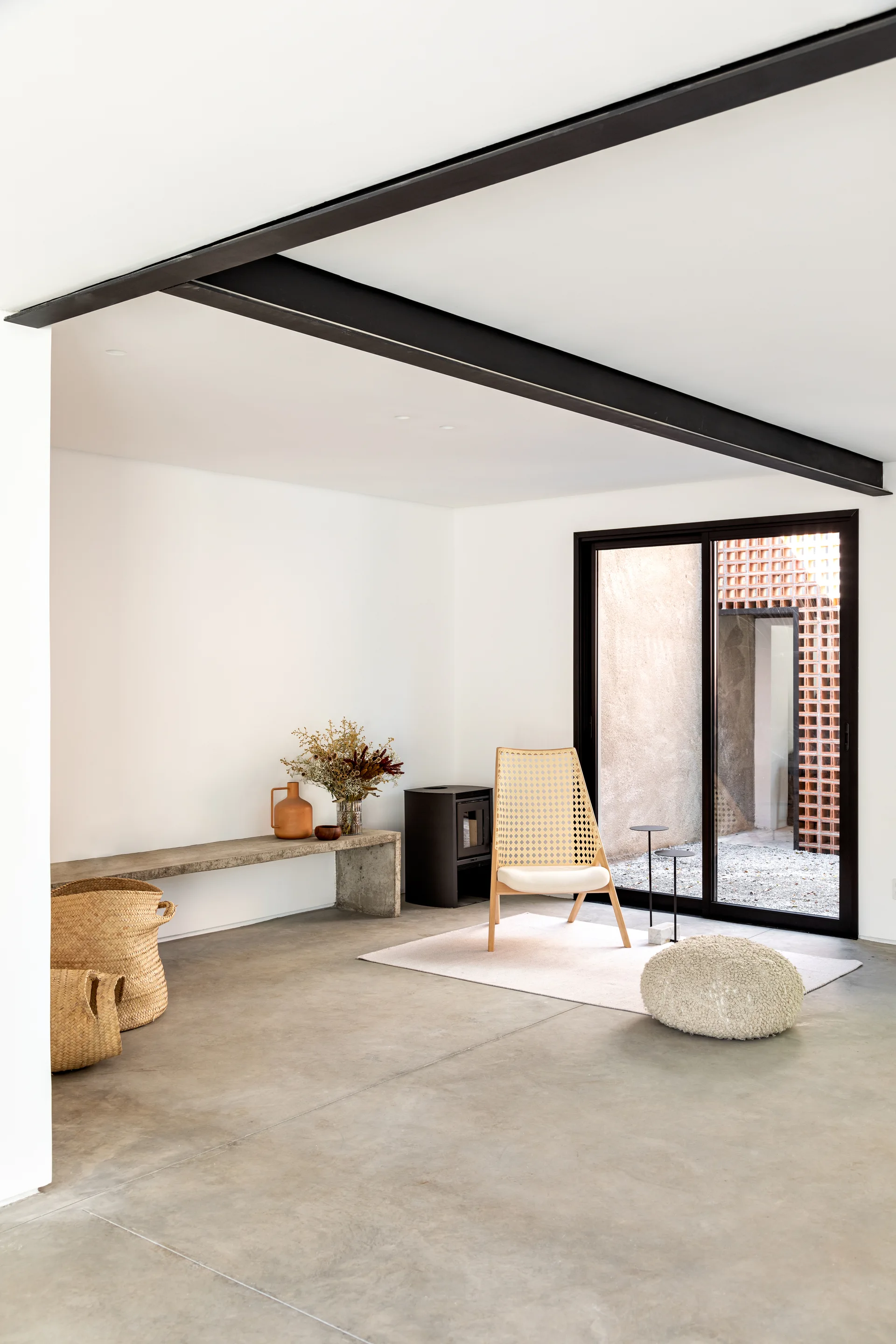 A minimalist living room features a cane lounge chair, a low concrete bench, and a black structural beam above polished concrete floors.
