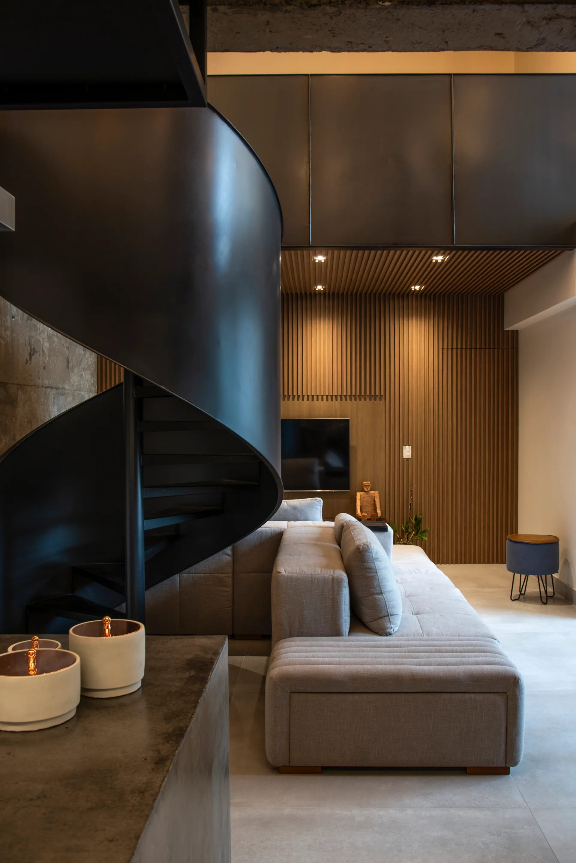 A modern living room featuring a prominent black metal spiral staircase, a grey sectional sofa, and a vertical wood slat accent wall.