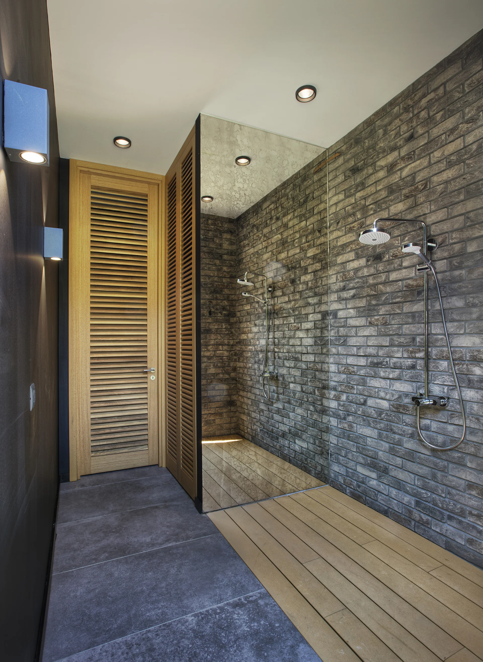 A modern bathroom featuring an exposed grey brick shower wall, chrome fixtures, a frameless glass partition, and a louvered wooden door.