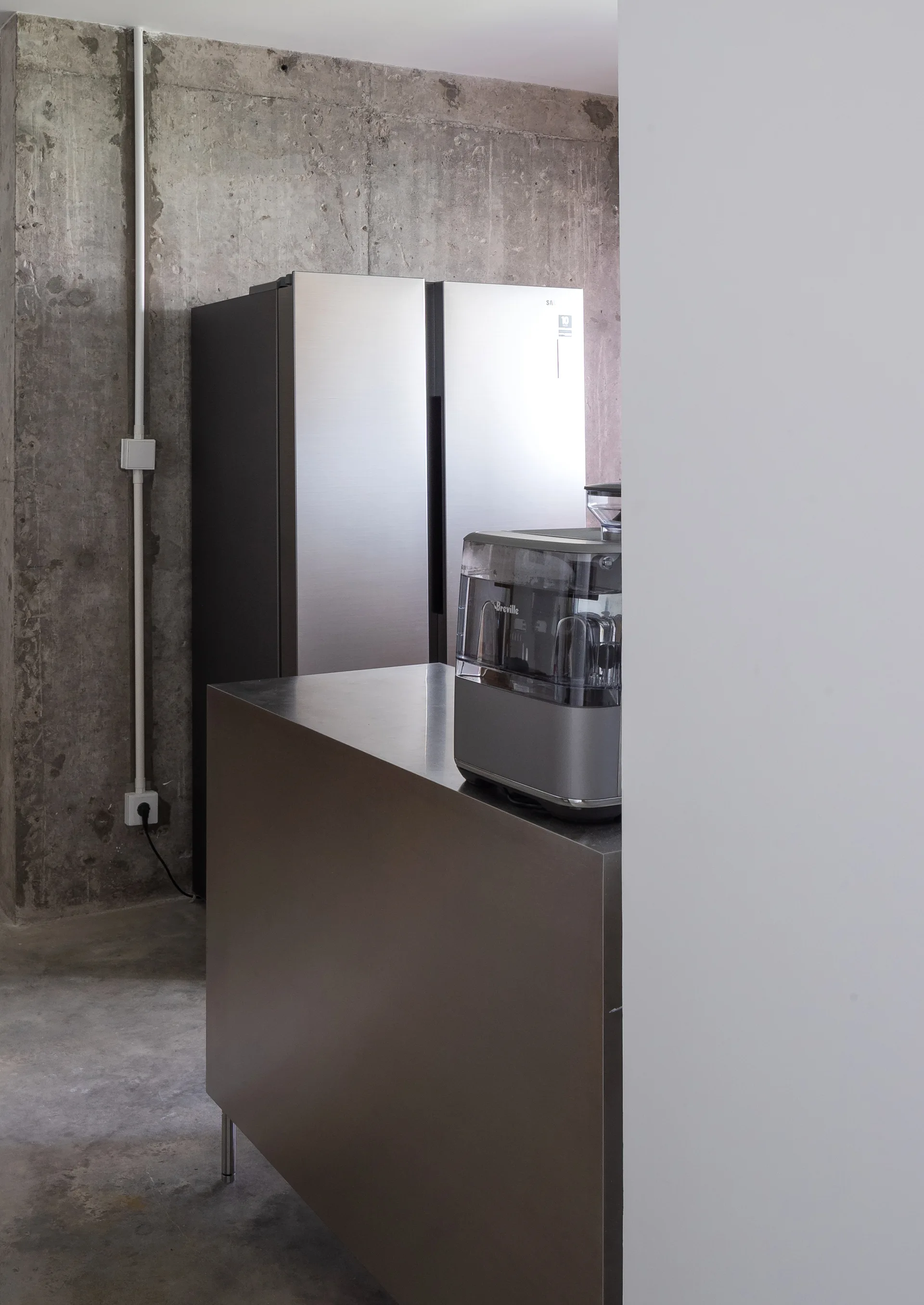 An industrial kitchen features a stainless steel refrigerator and island against a raw exposed concrete wall with a white surface-mounted conduit.