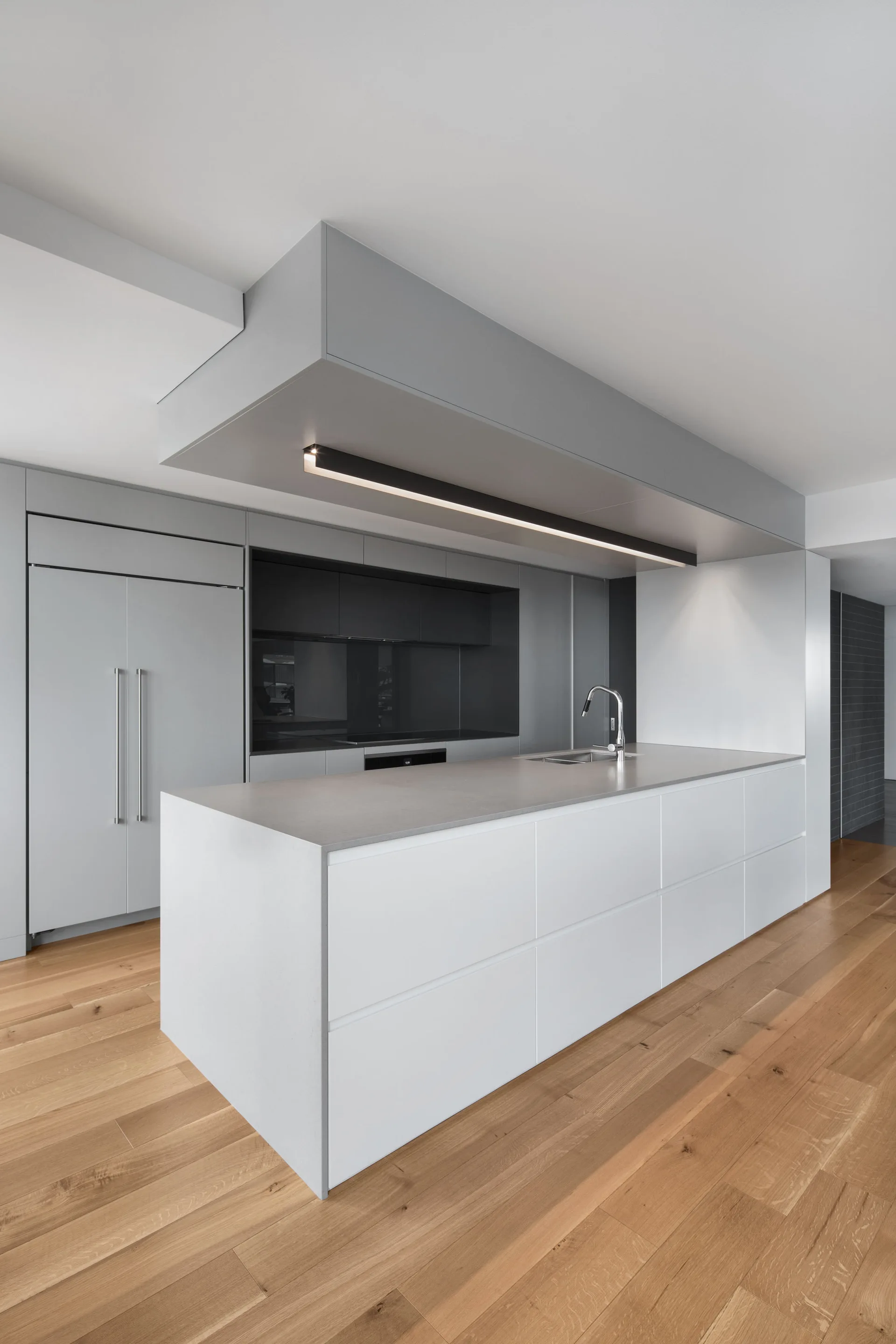 A minimalist kitchen features a white central island, integrated grey cabinets, a suspended ceiling soffit with a linear light, and light wood floors.