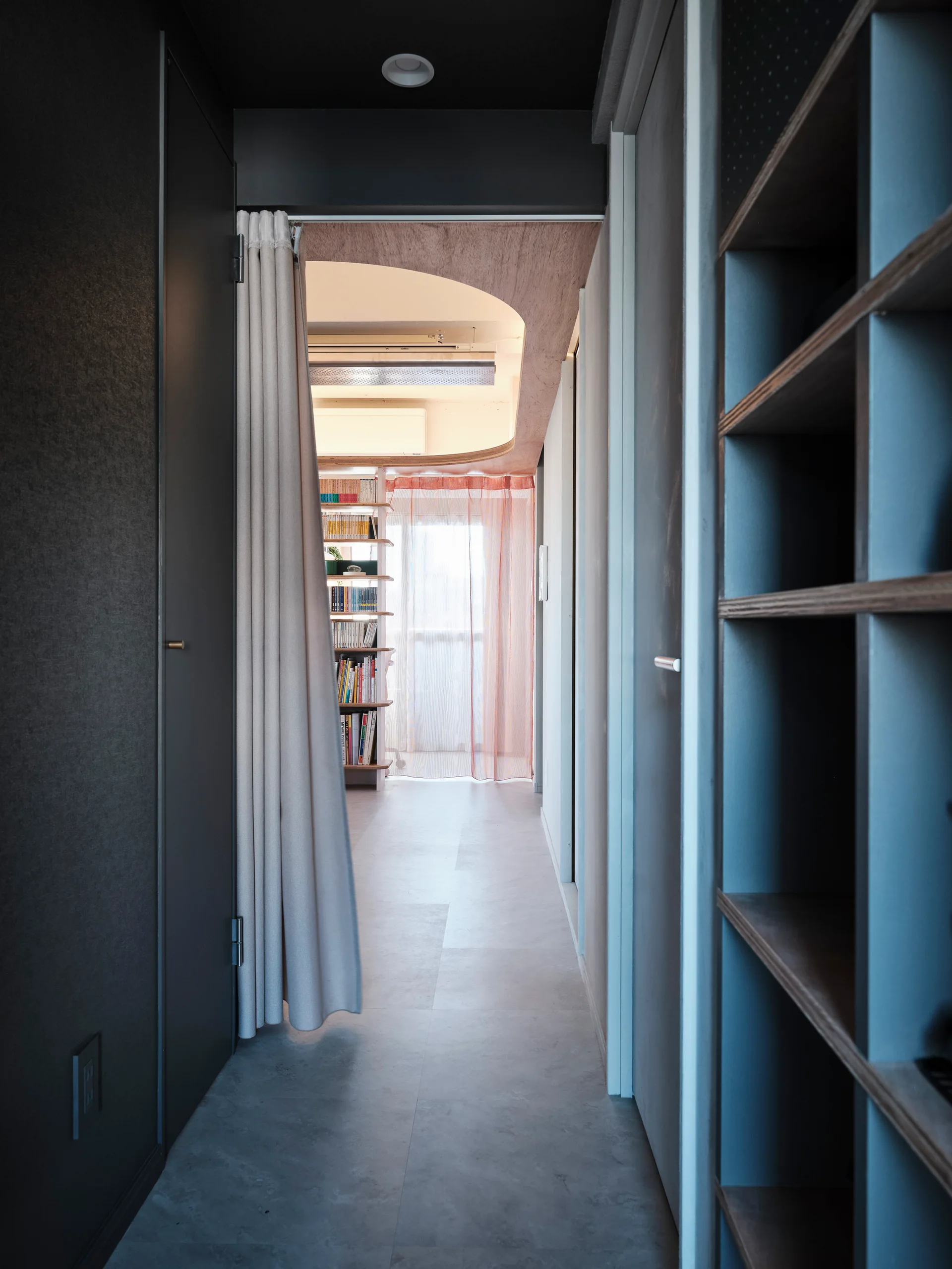 A grey residential hallway featuring built-in shelving leads through an arched ceiling transition to a sunlit room with pink sheer curtains.