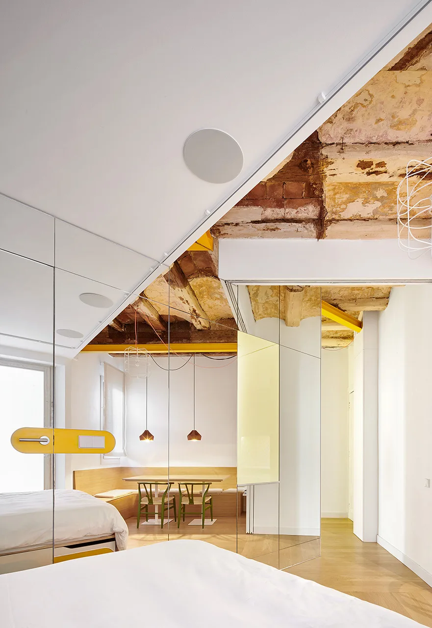 A modern bedroom features a white bed next to a large mirrored wall partition reflecting a dining nook beneath rustic, exposed ceiling beams.