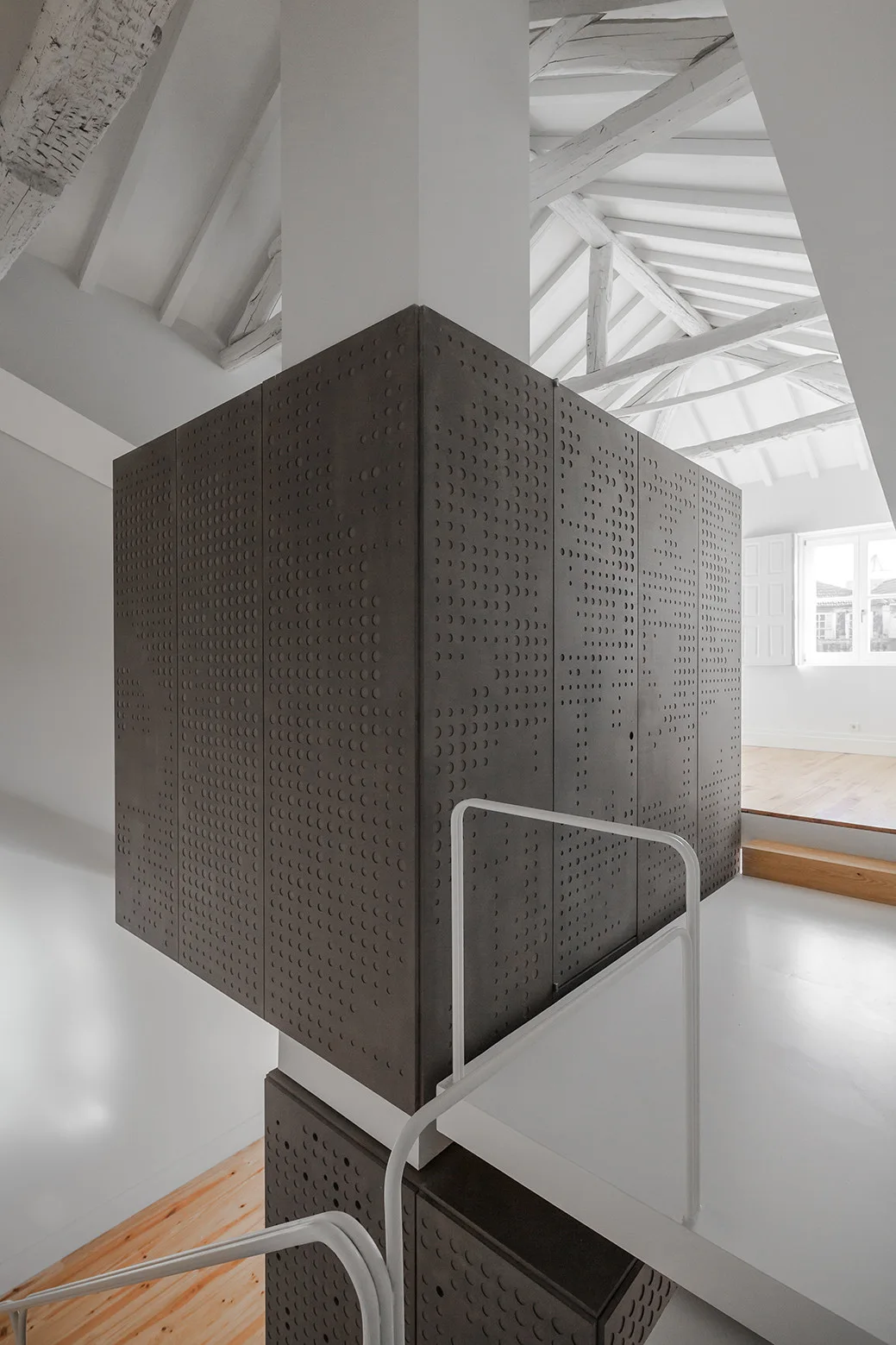 A dark grey perforated partition block stands in a minimalist white loft with a vaulted beamed ceiling and white staircase handrails.