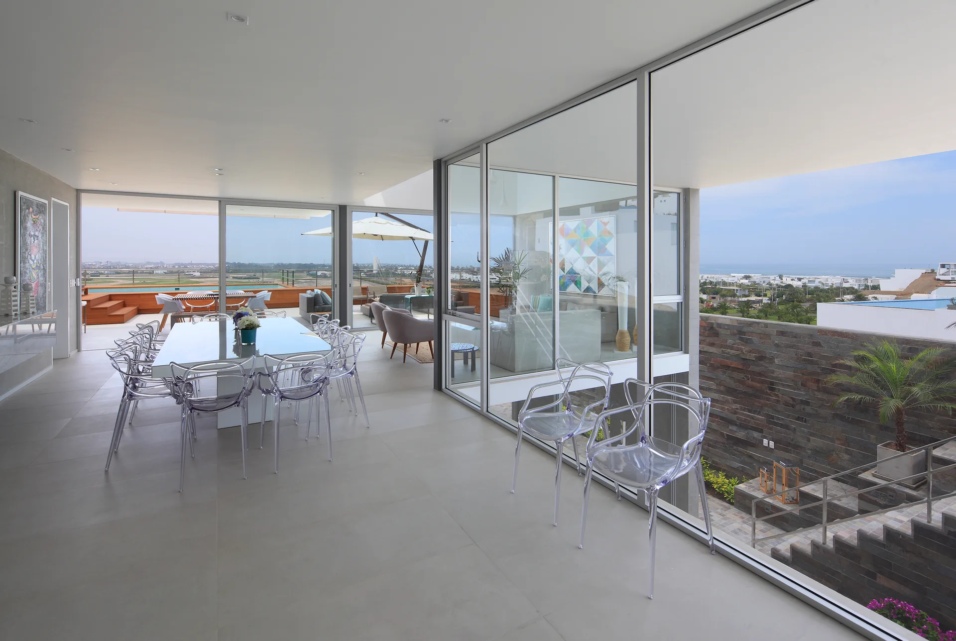 A modern indoor dining room features a white table with clear ghost chairs next to expansive glass doors that open to an outdoor terrace.