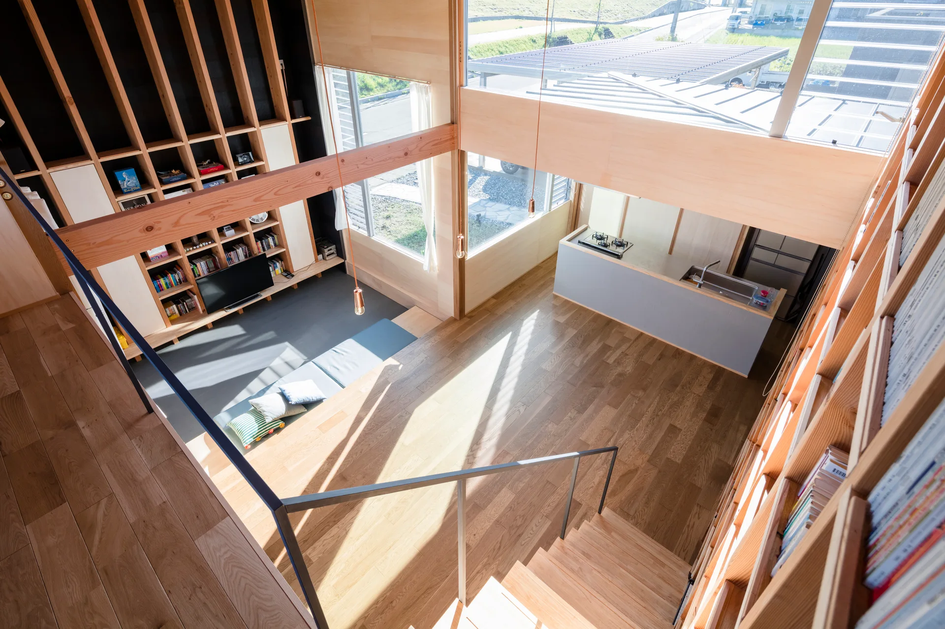A high-angle view of a modern open-plan home featuring a massive light wood bookshelf wall, a wooden staircase, and a minimalist kitchen counter.