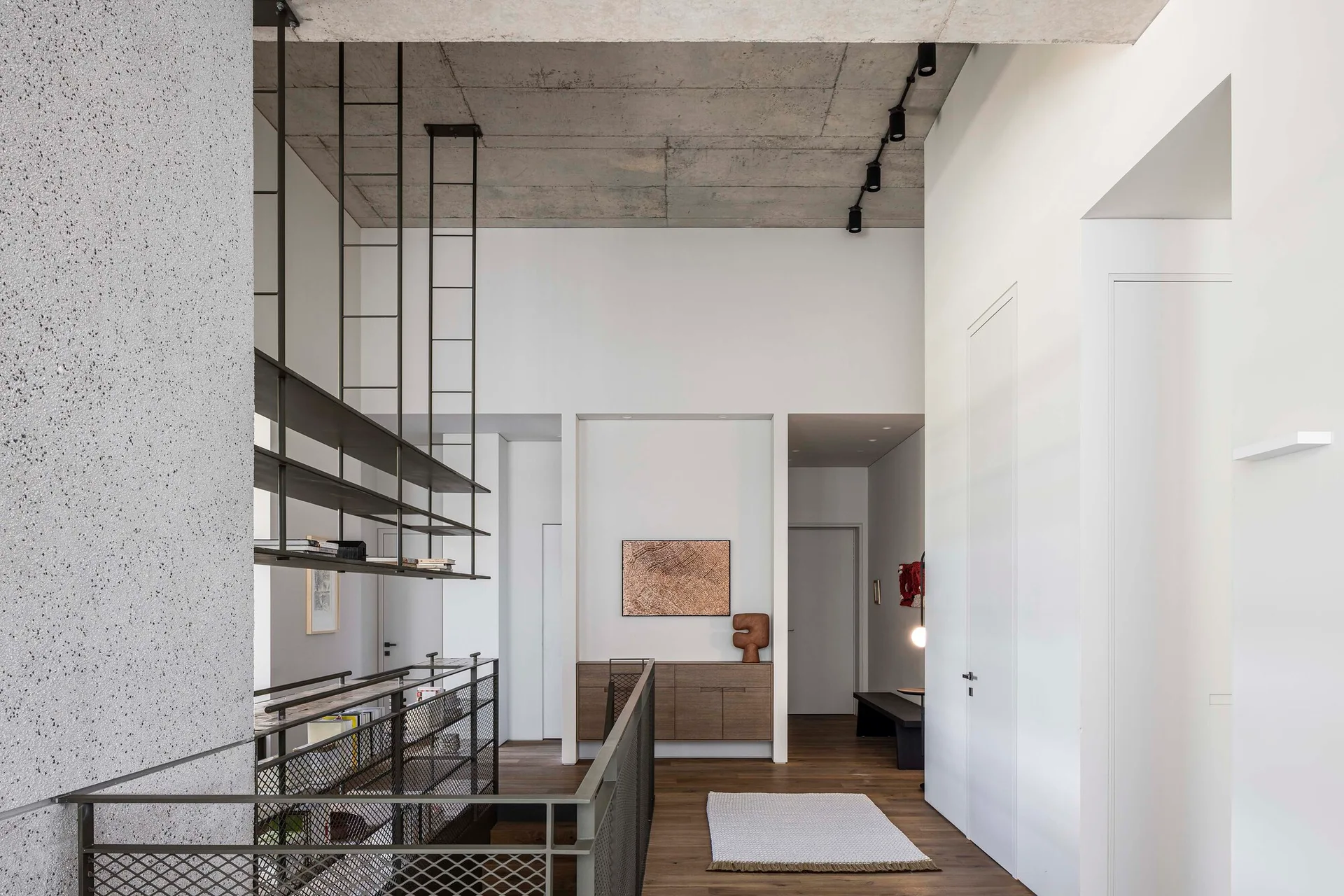 A minimalist staircase landing features an exposed concrete ceiling, suspended black metal shelves, a wire mesh railing, and a low wooden console.