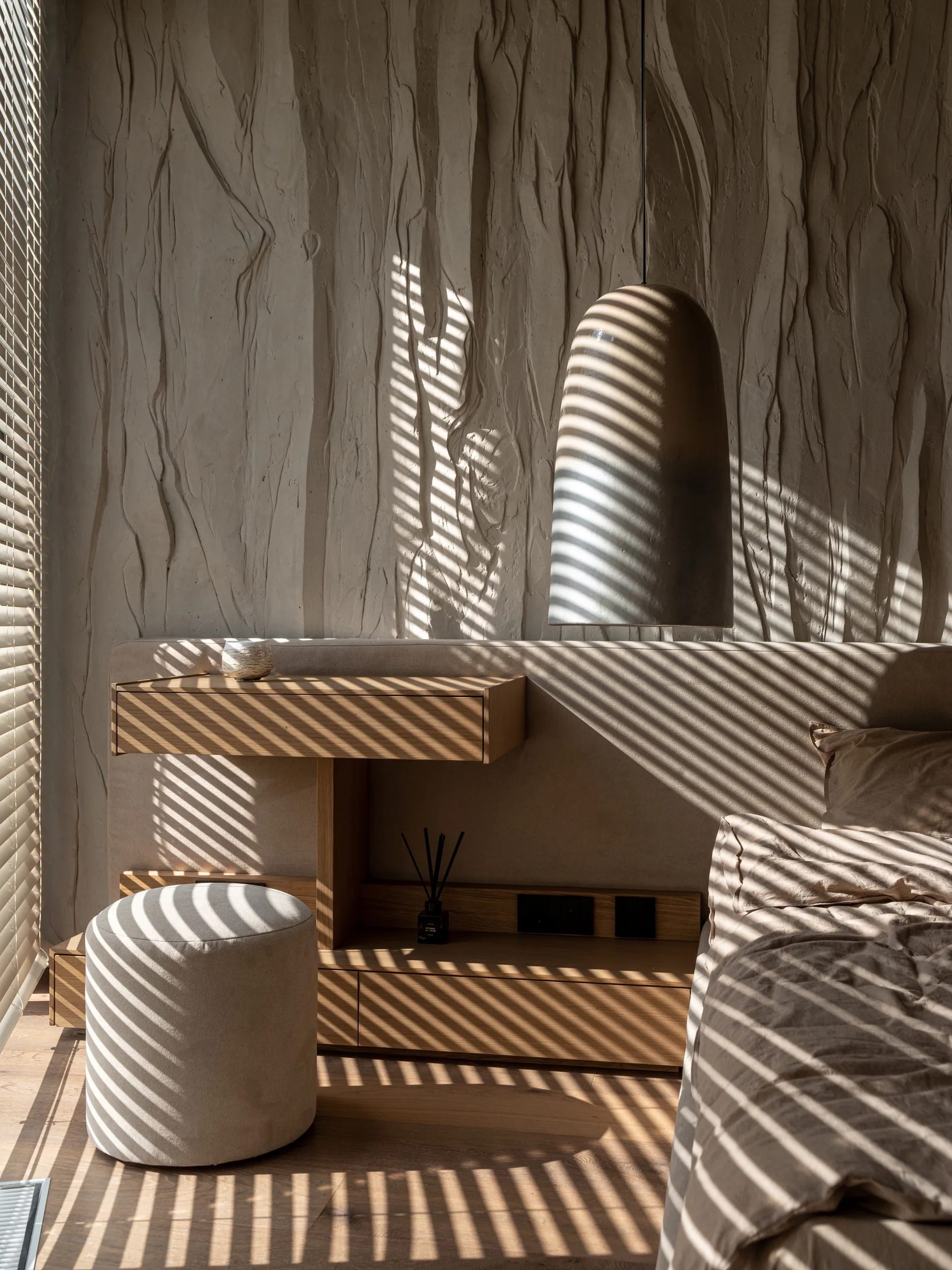 A minimalist bedroom features an integrated wooden desk and headboard against a textured plaster wall, with slatted blinds casting linear shadows.
