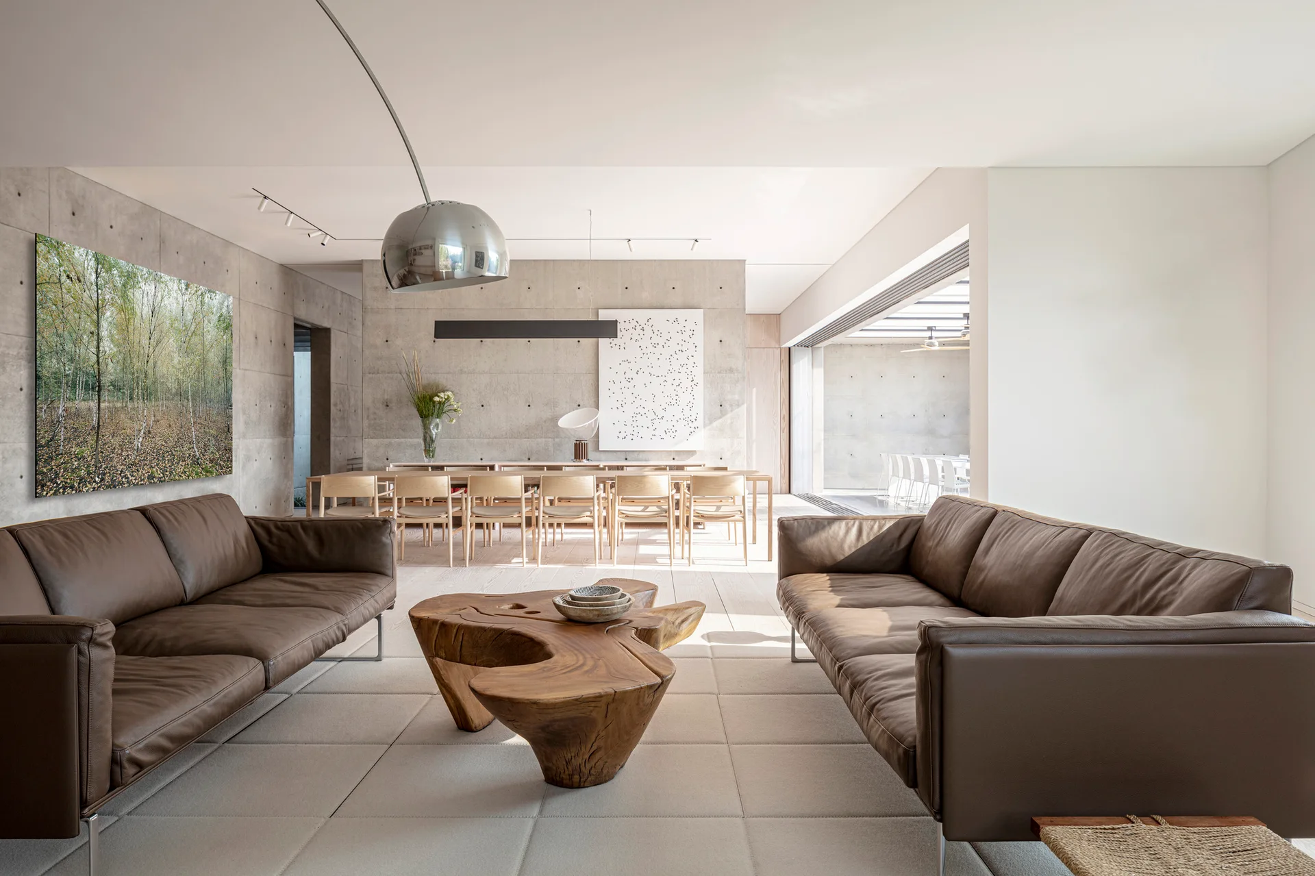 An open living space features brown leather sofas, a sculptural wood coffee table, and exposed concrete walls with a dining area in the background.