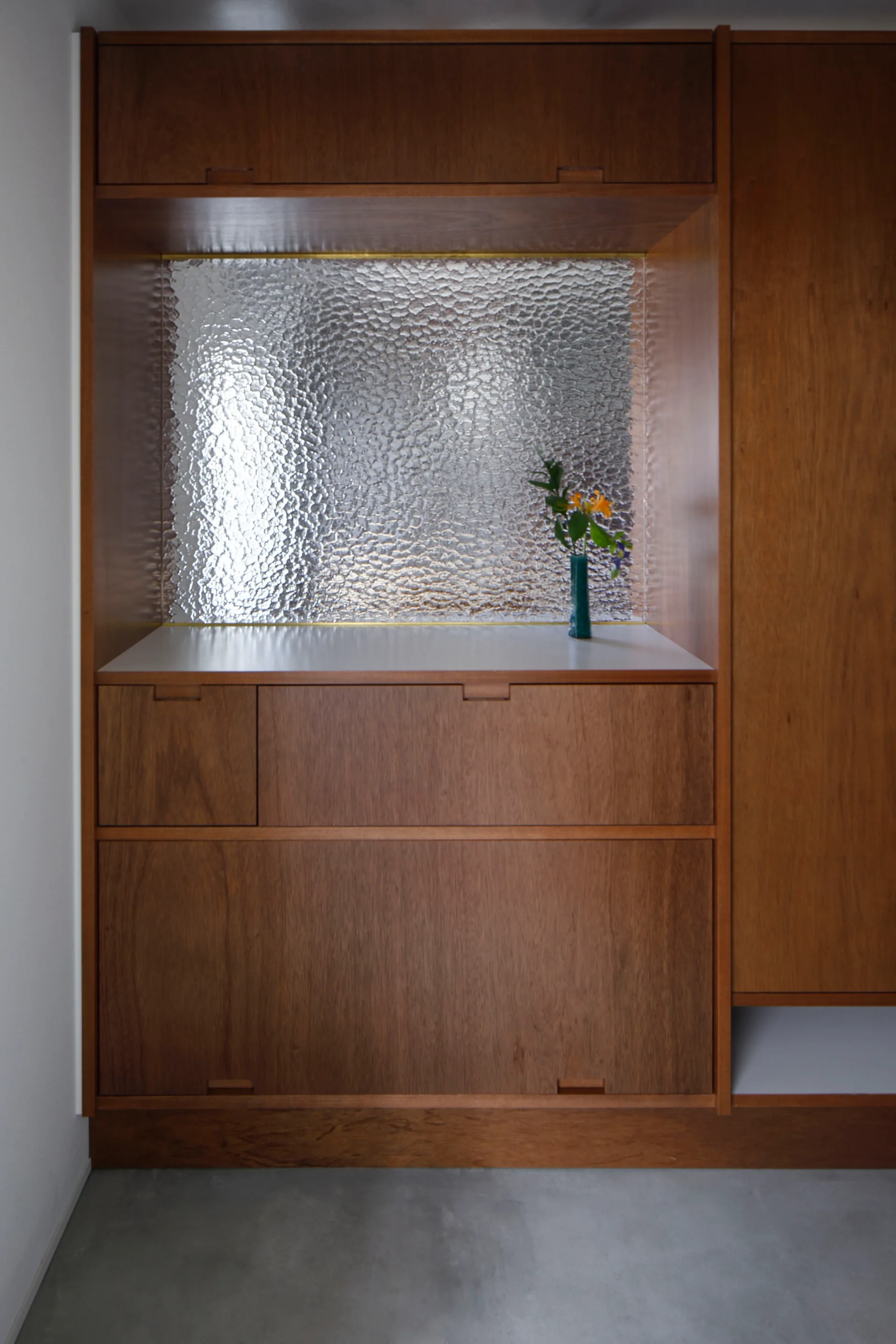 A minimalist wood built-in cabinet featuring a recessed countertop with a textured silver backsplash and a small green flower vase.