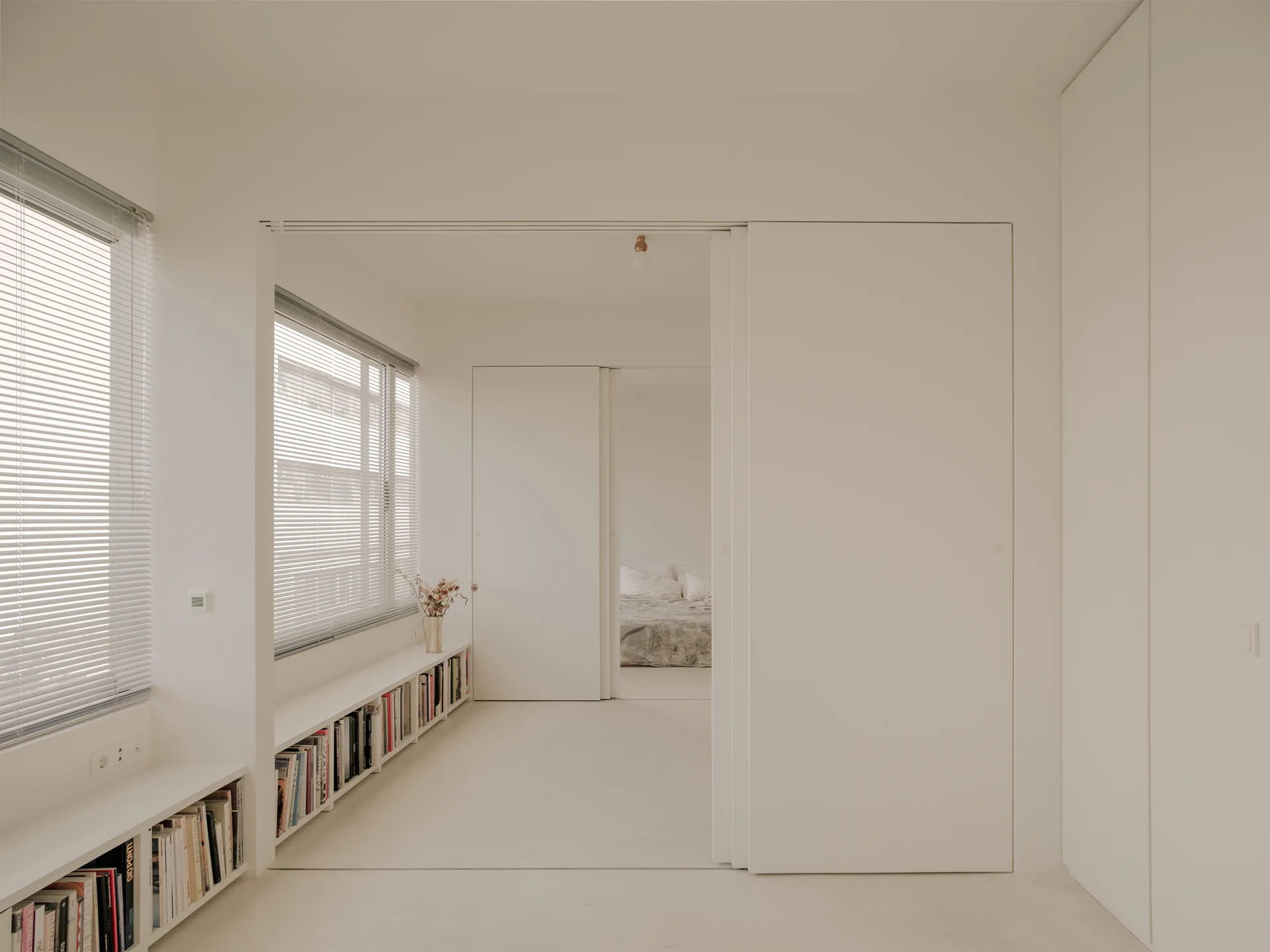 A minimalist white room features low built-in bookshelves beneath windows and large sliding doors that open into a bedroom.
