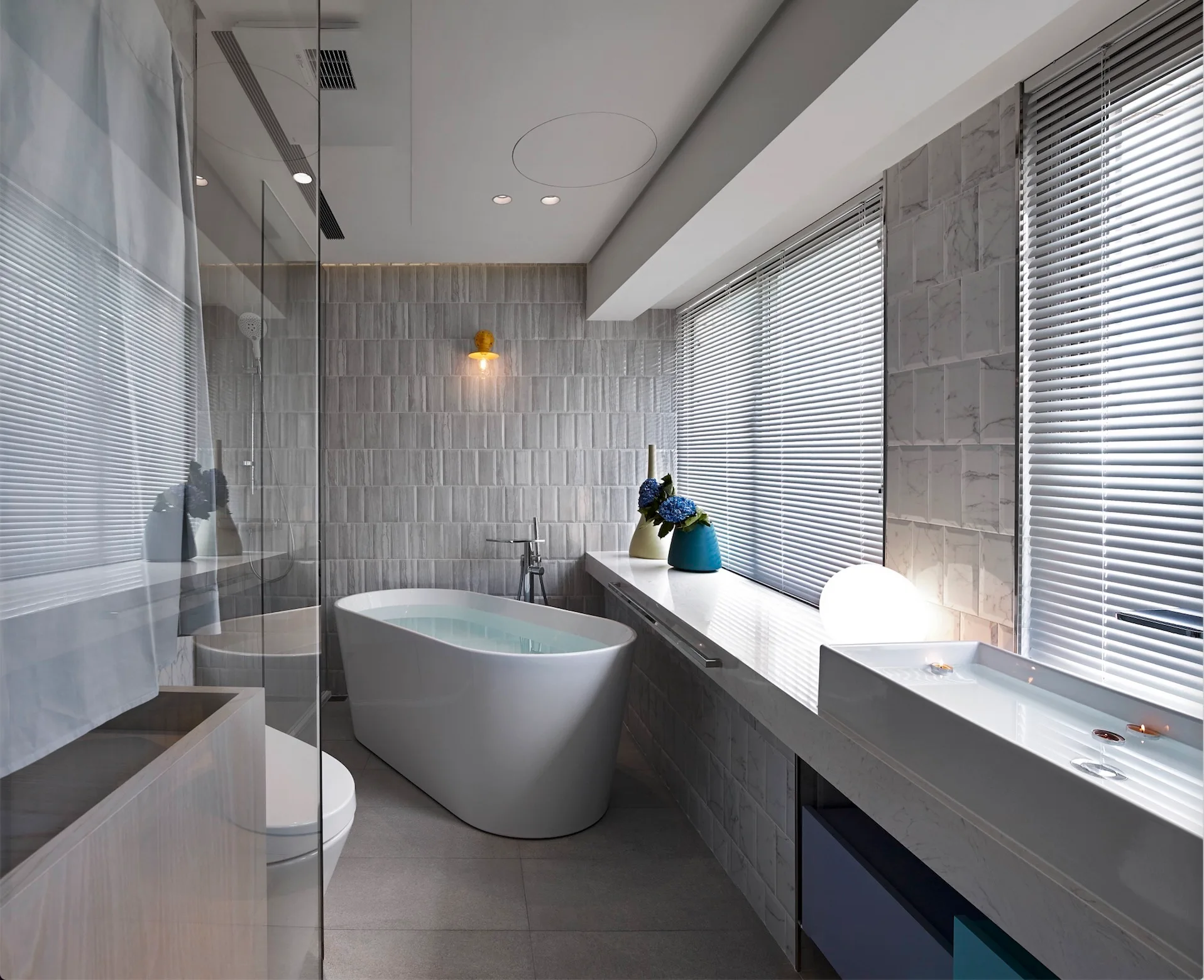 A modern gray bathroom features a white freestanding bathtub, a long polished counter with a sink, and window blinds.