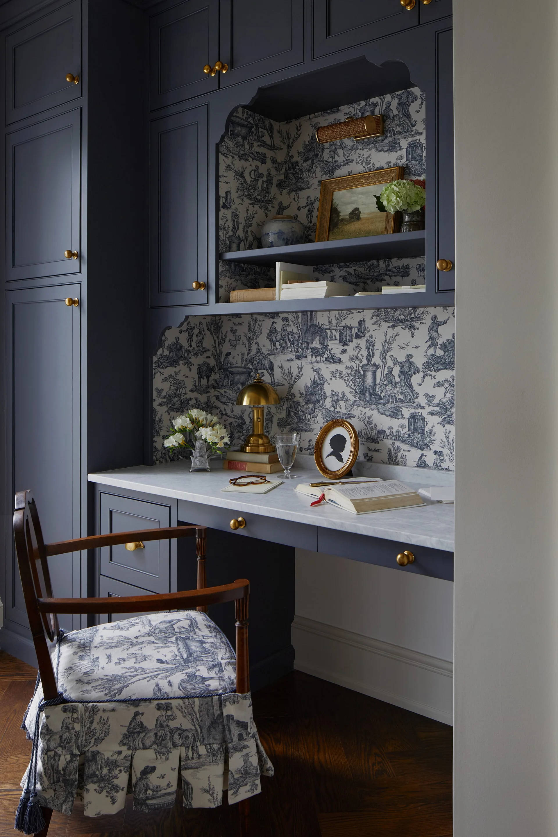A dark blue built-in desk nook features a white marble desktop, blue and white toile wallpaper, and a matching upholstered wooden armchair.