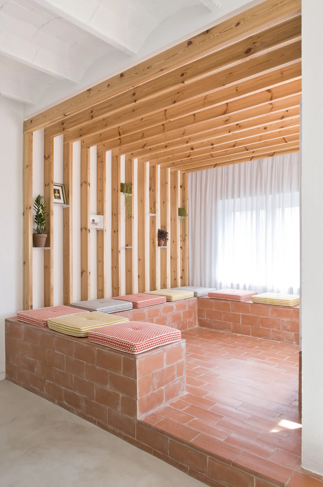 A built-in terracotta brick bench with patterned cushions sits beneath a natural wood slat wall and ceiling canopy in a bright room.