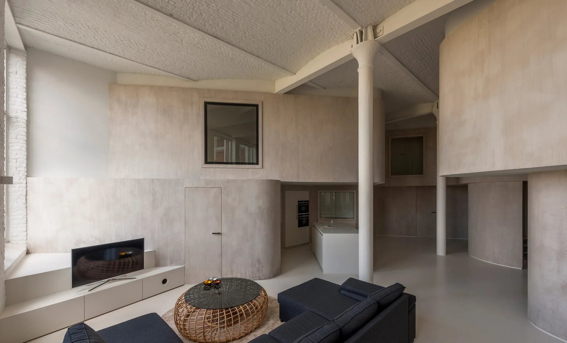 An open loft apartment features curved plaster walls, a dark grey sectional sofa, a round wicker coffee table, and white structural columns.
