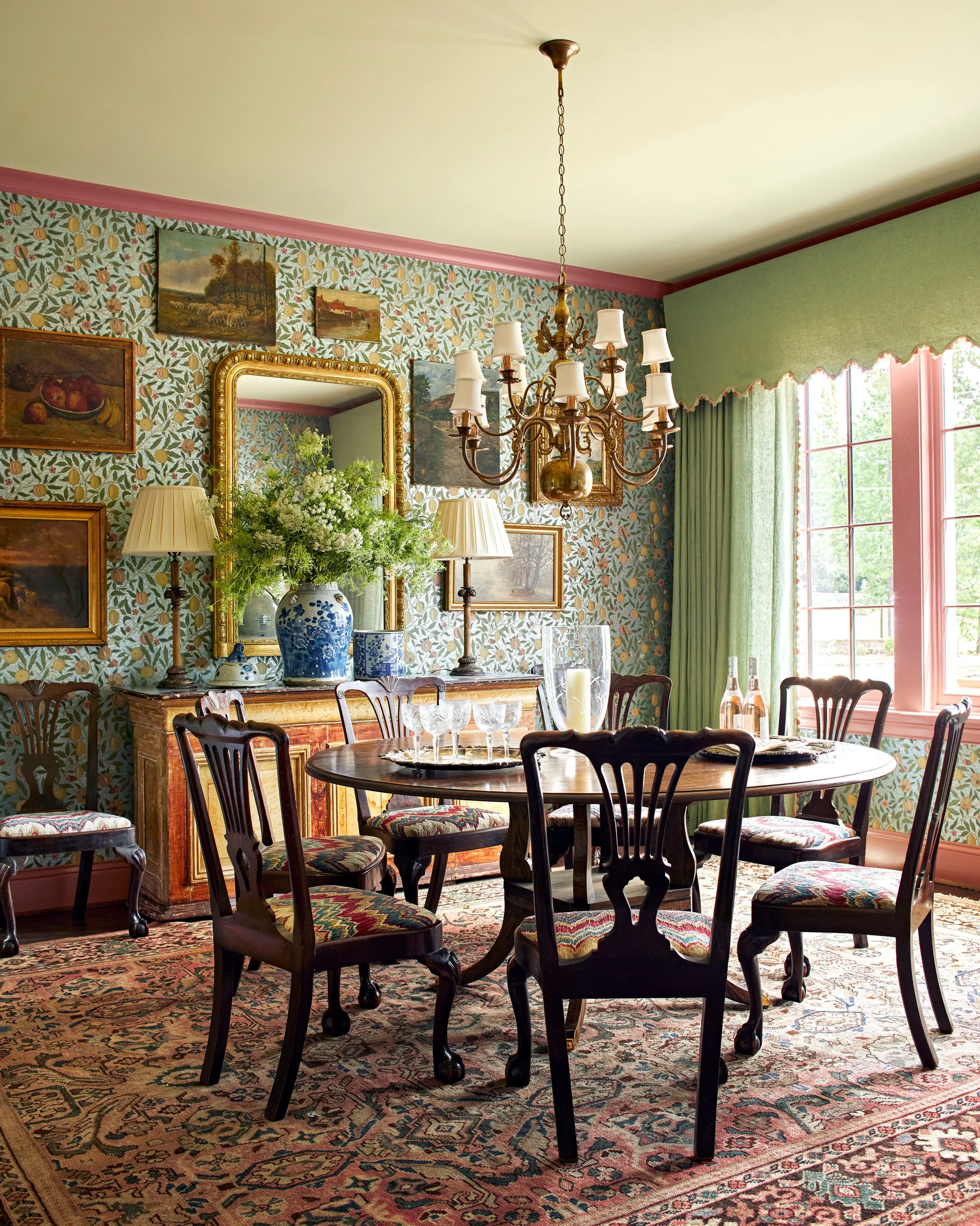A traditional dining room featuring a round wooden table, chairs with patterned seats, botanical wallpaper, and a gold-framed mirror.