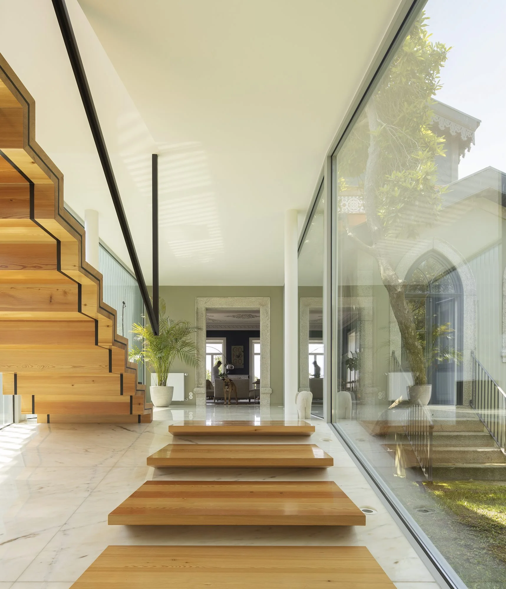 A bright hallway features floating wooden stairs on a polished marble floor next to a large glass wall looking out at a courtyard garden.