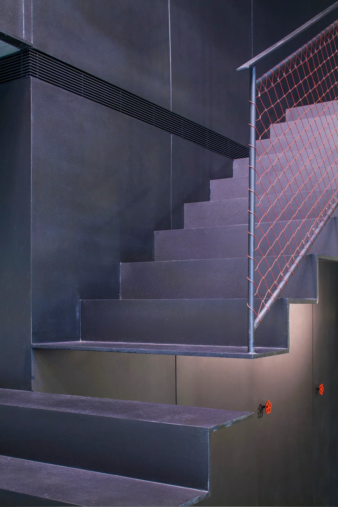 A matte black metal staircase rises against matching dark wall panels, accented by a red safety net attached to the handrail.