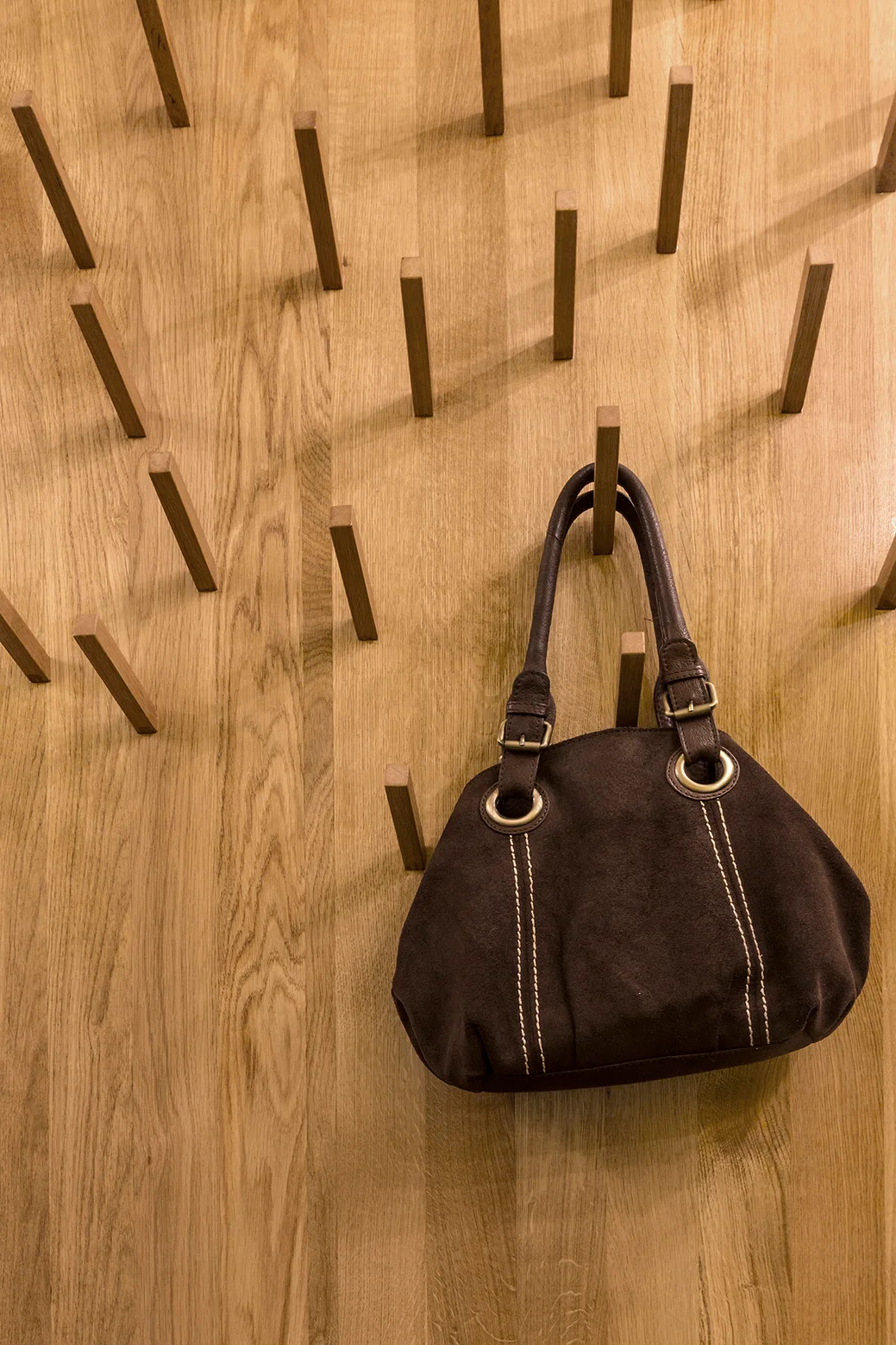 A brown suede handbag hangs from a light oak wood peg wall display with multiple projecting pegs.