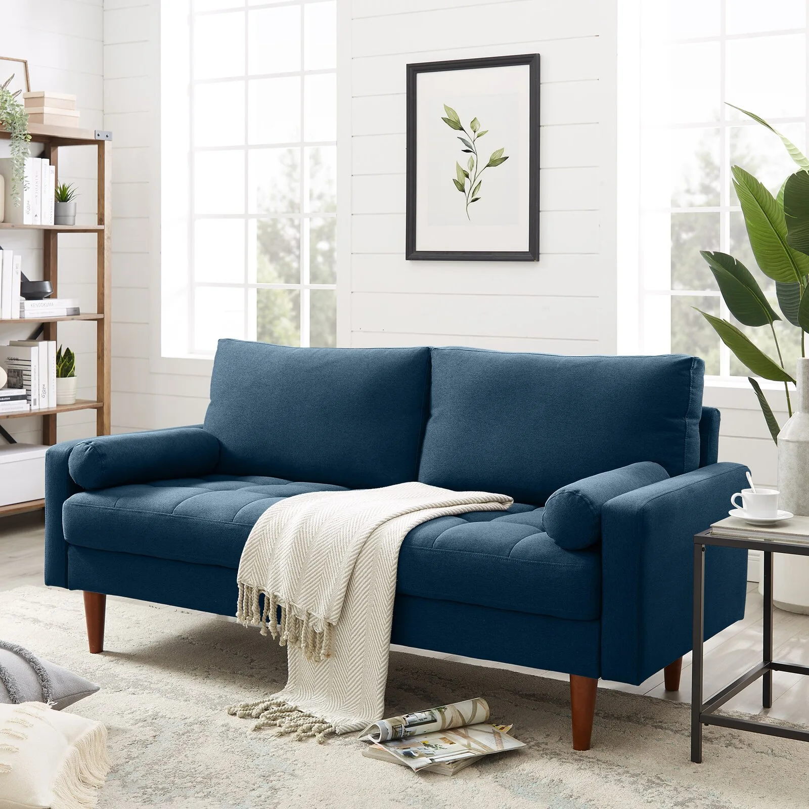 A deep blue upholstered sofa with wooden legs sits in a bright room with white horizontal wood paneling, large windows, and a wood bookcase.