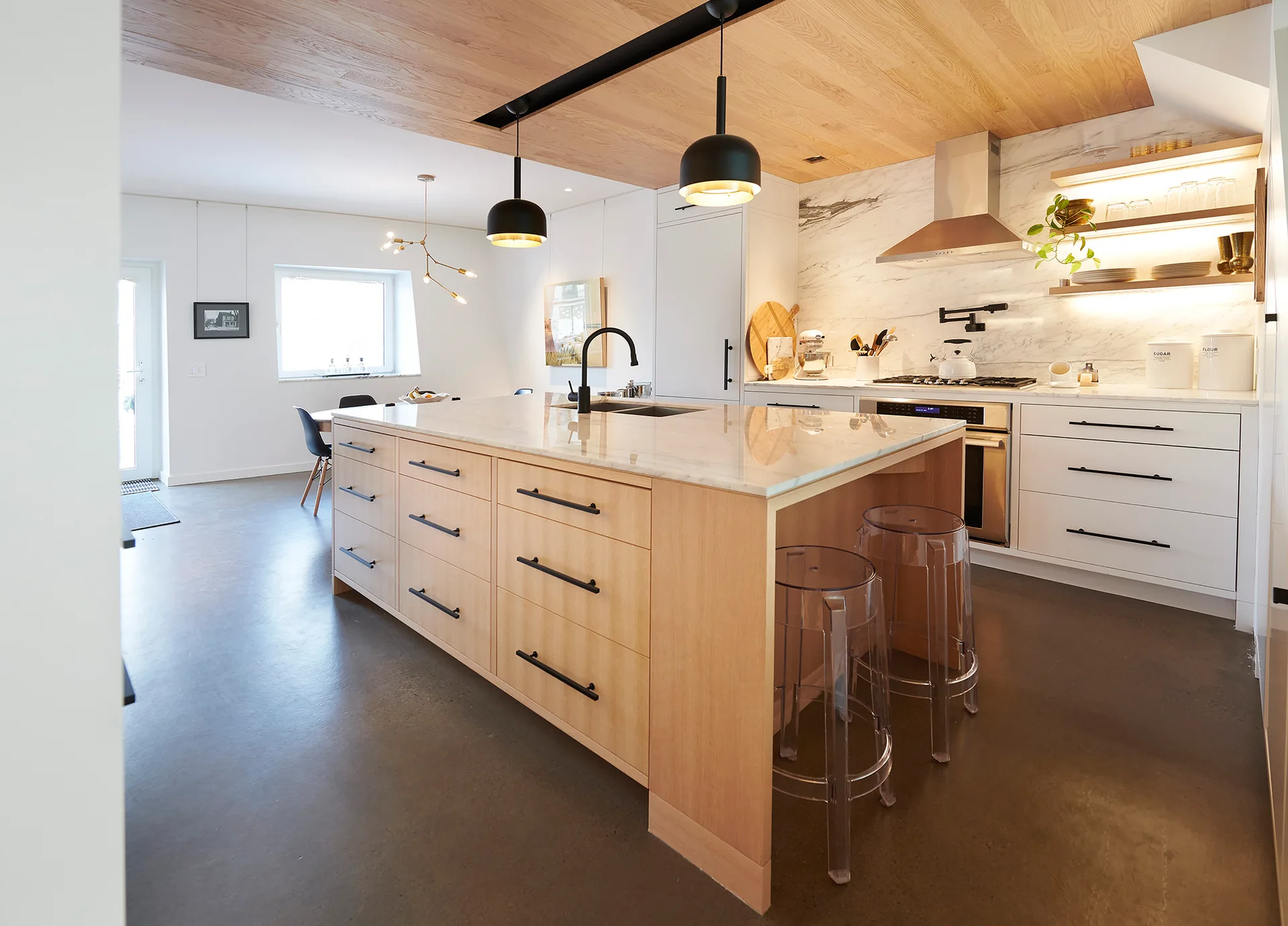 A modern kitchen features a light wood island with a marble countertop, white cabinets, clear stools, and a warm wood plank ceiling.