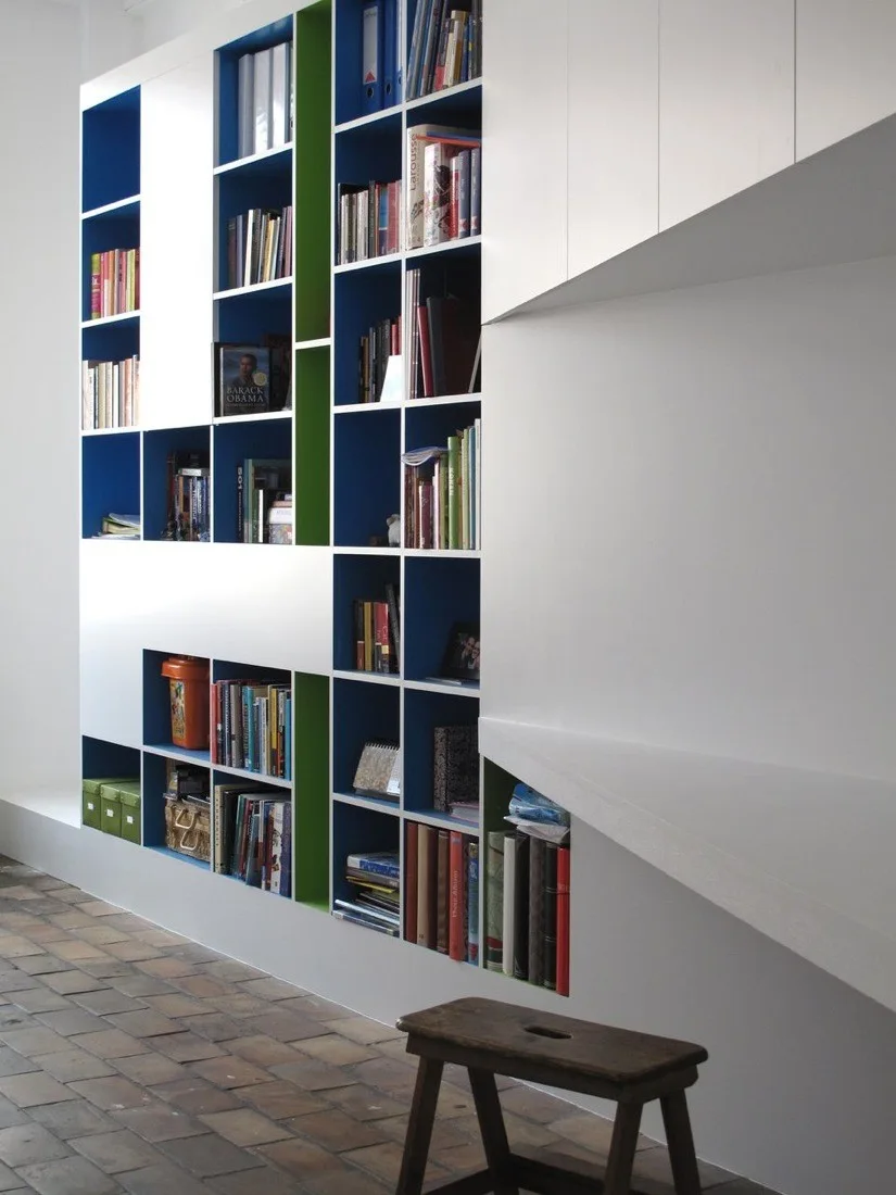 A modern white built-in shelving unit with bright blue and green inner cubbies filled with books above a terracotta tile floor.