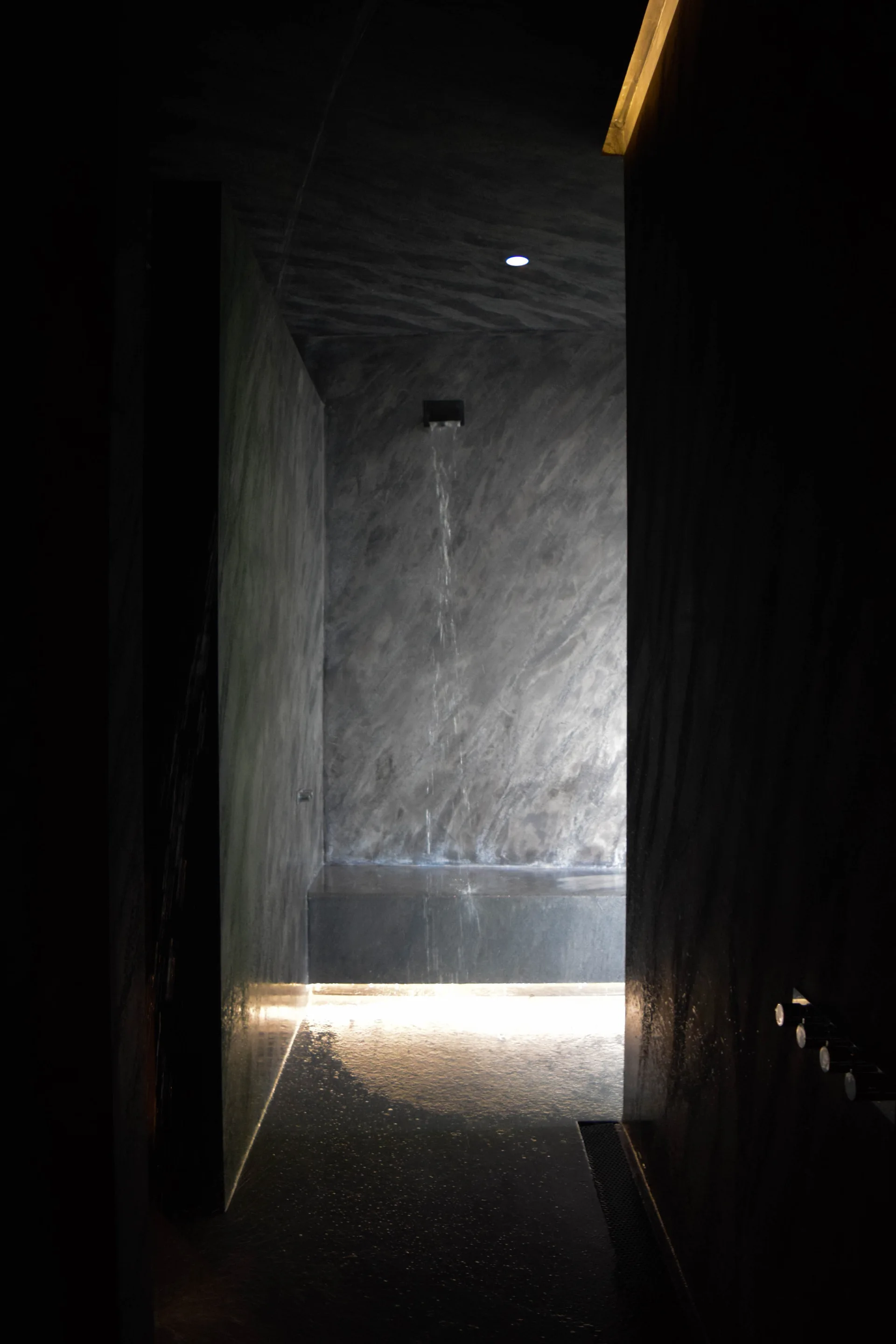 A minimalist charcoal stone shower stall features water flowing from a wall fixture and warm glowing light beneath a built-in bench.
