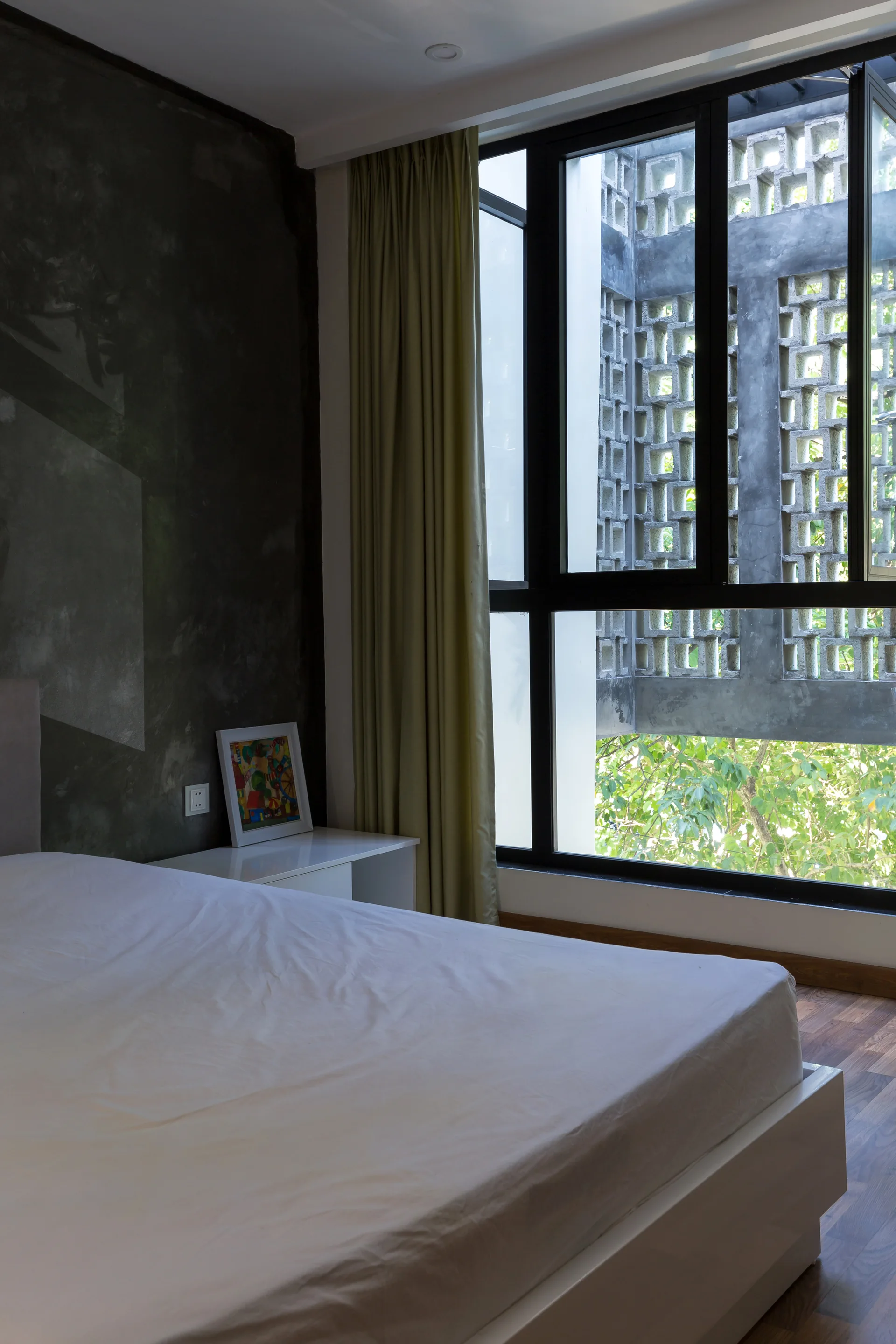 A modern bedroom with a white bed, a dark textured concrete wall, and a large window looking out at a patterned concrete breezeblock screen.