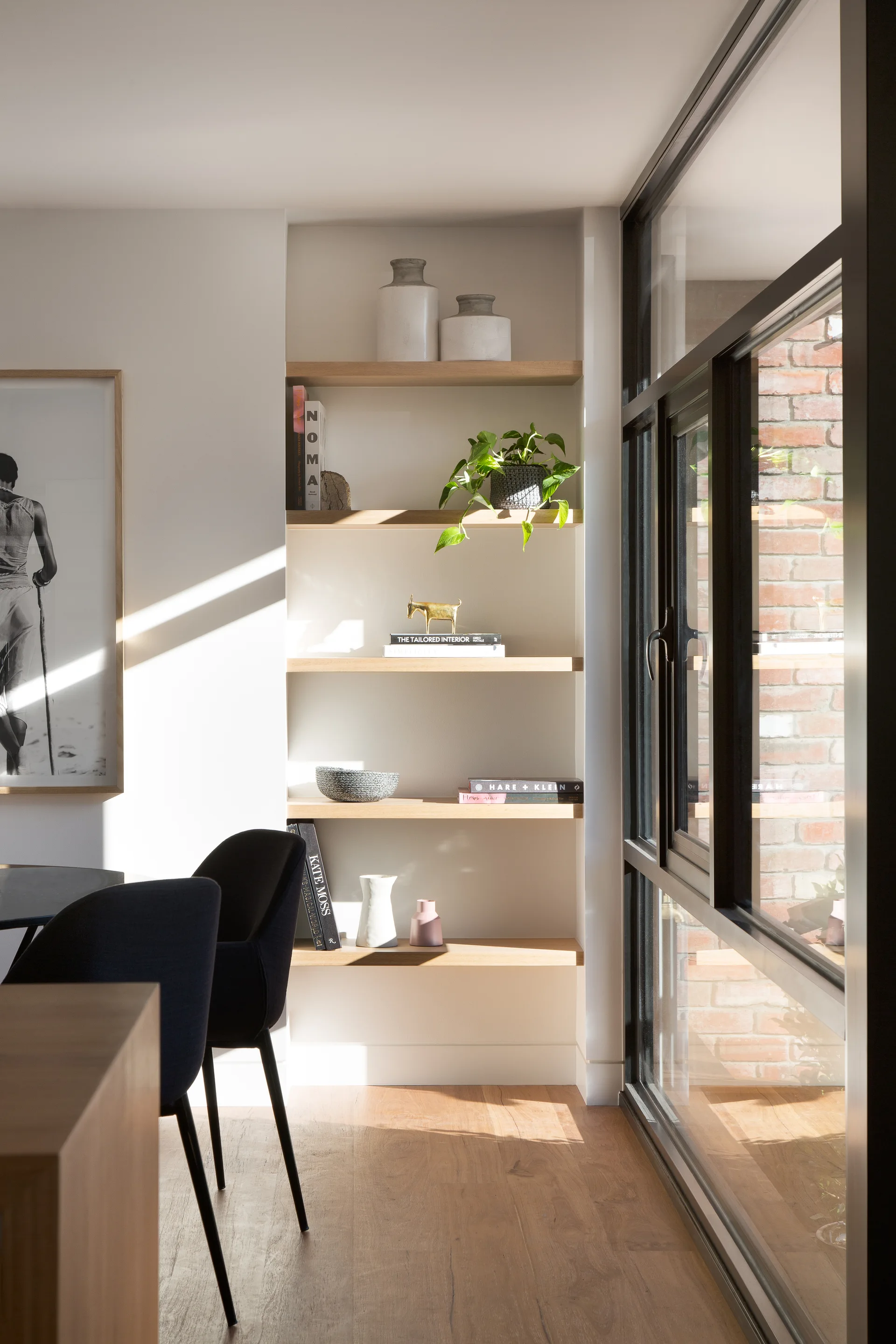 Sunlight shines through a large black-framed window onto recessed oak shelves decorated with books, vases, and a small plant.