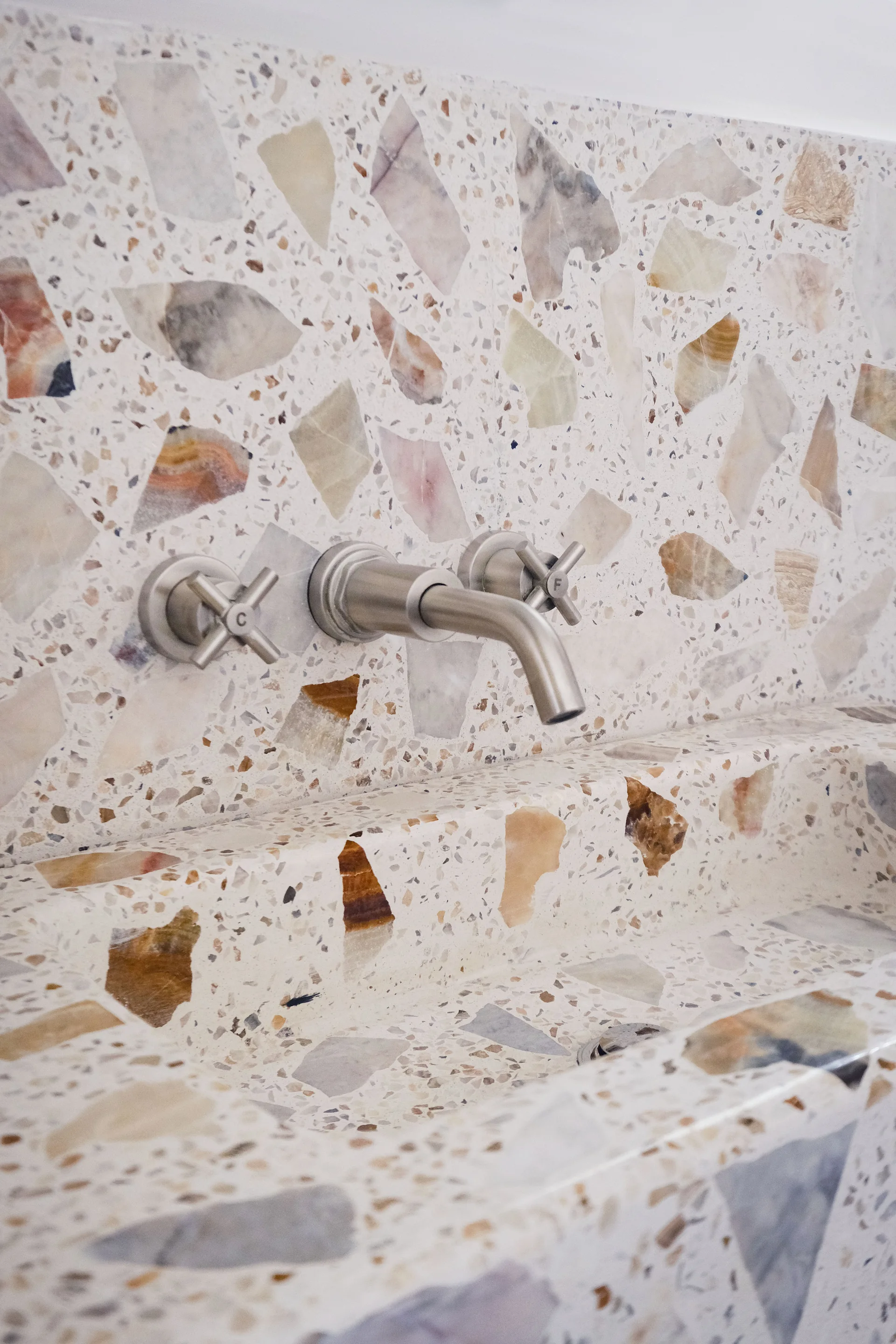 A brushed metal wall-mounted faucet with cross handles is set against a terrazzo backsplash and integrated stone sink basin.