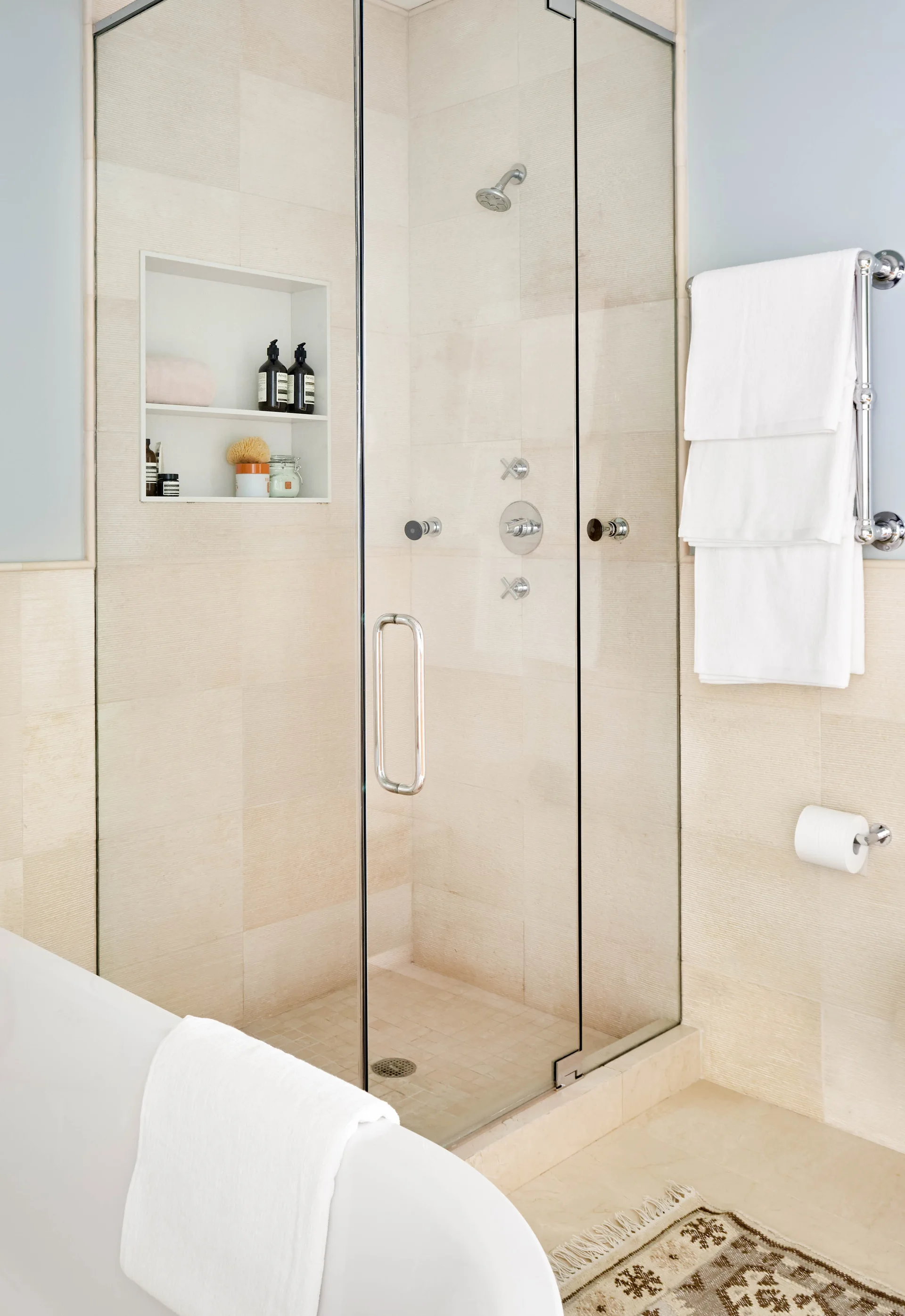 A glass shower enclosure with cream textured wall tiles, a built-in storage niche, chrome fixtures, and white towels hanging on a nearby rack.