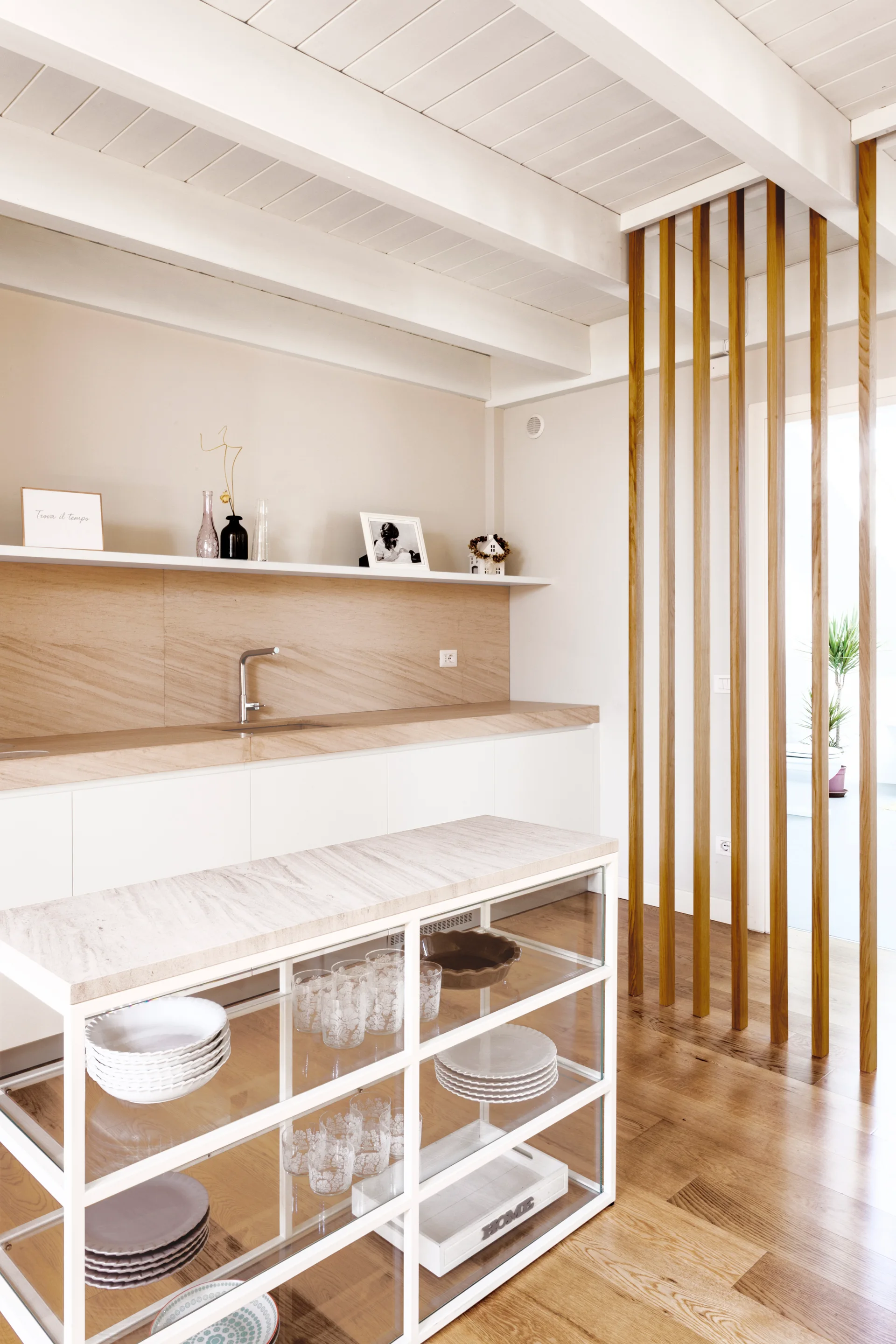 A modern kitchen featuring a glass-shelved island, a wooden slat room divider, beige stone countertops, and white painted ceiling beams.