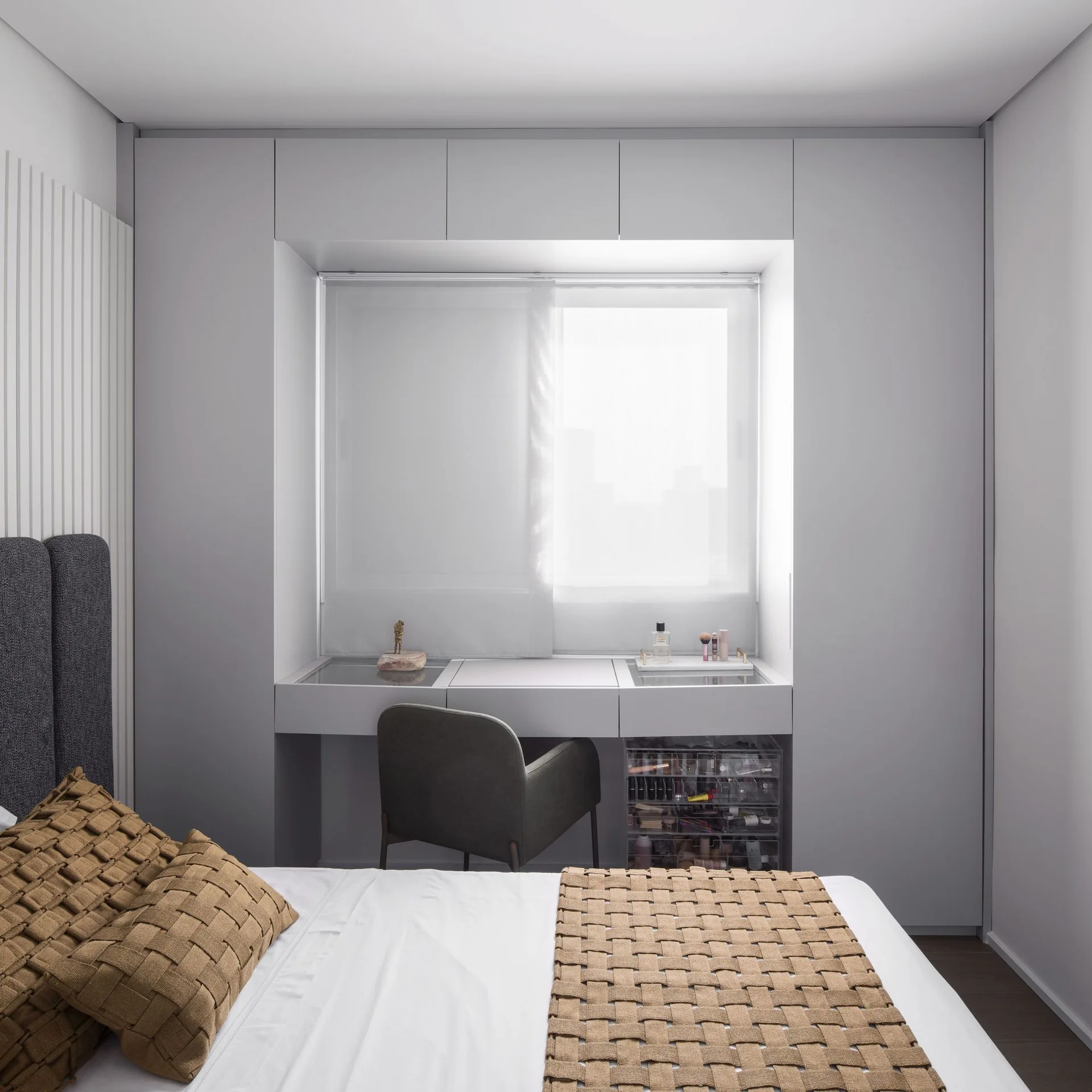 A modern bedroom features a light gray built-in wardrobe framing a window with an integrated vanity desk, a gray chair, and woven tan bedding.