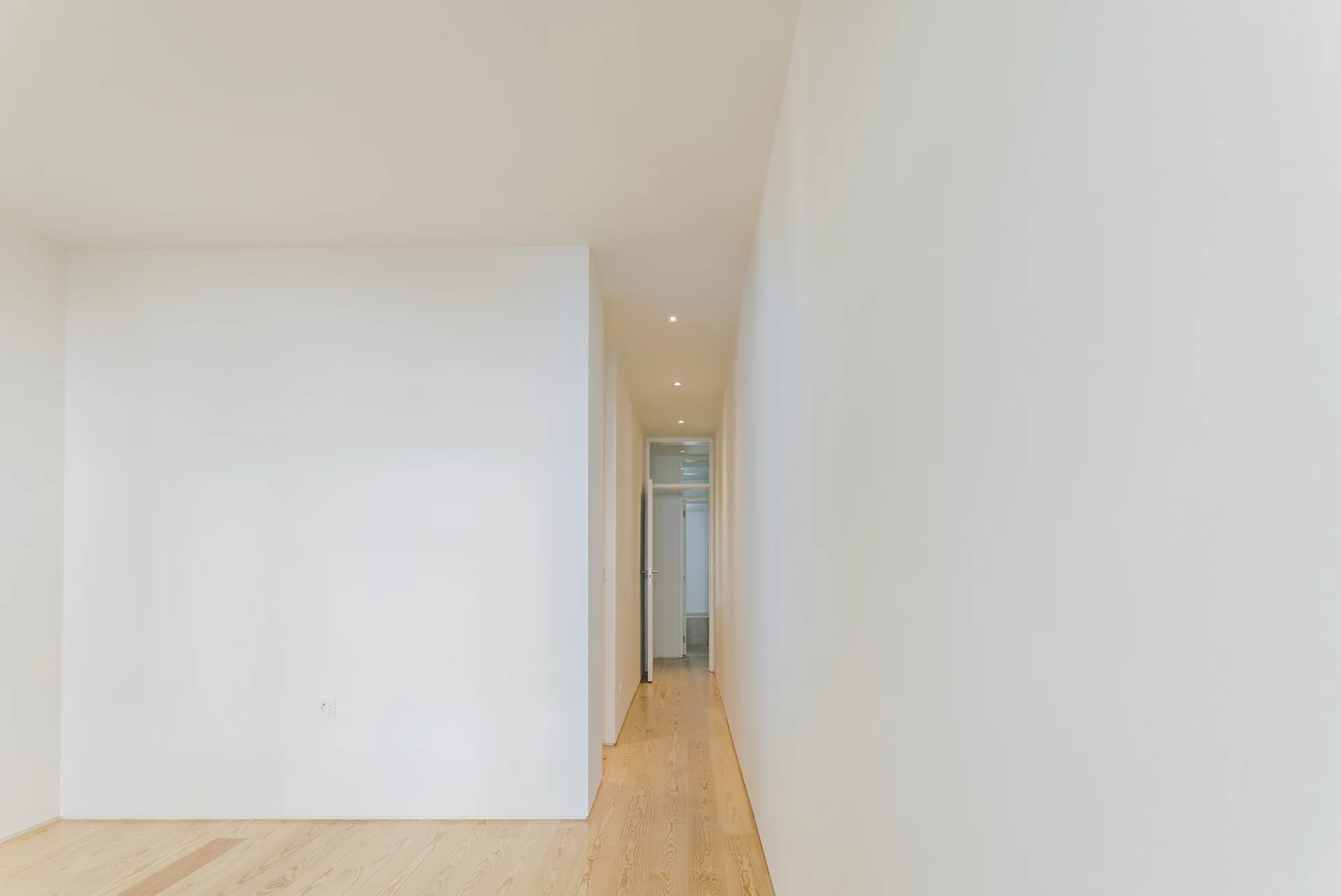 A long minimalist hallway features plain white walls, light wood plank flooring, and small recessed ceiling lights leading to an open doorway.