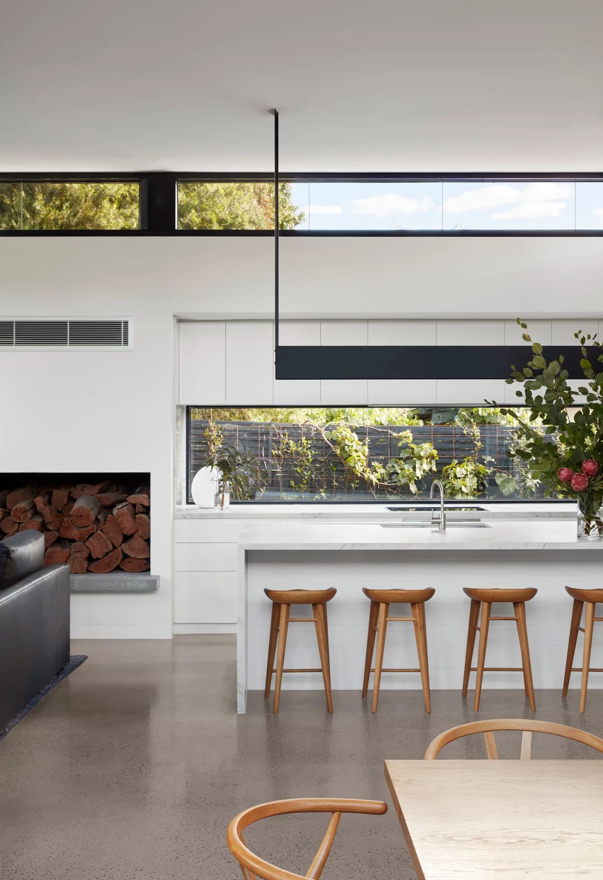 A modern white kitchen features a marble-topped island with wooden stools, high clerestory windows, and a built-in niche filled with stacked logs.