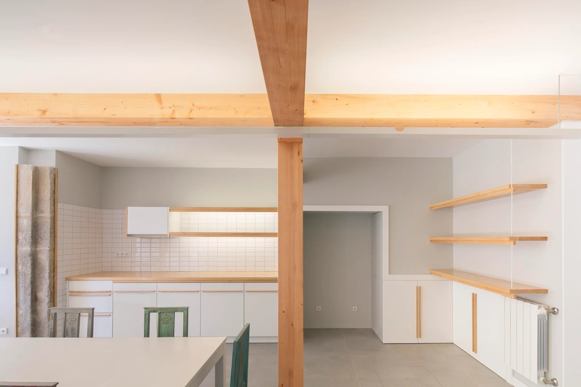 A minimalist kitchen features white cabinetry, light wood countertops, open shelving, and a prominent vertical timber support post.