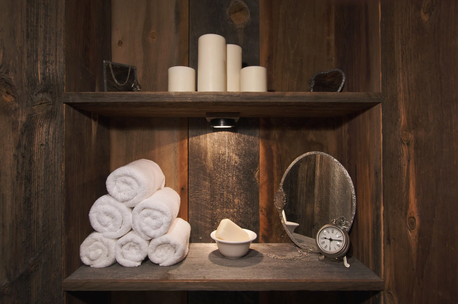 A rustic wood shelving unit built into a plank wall holds a stack of rolled white towels, a small vintage mirror, and pillar candles under a spotlight.