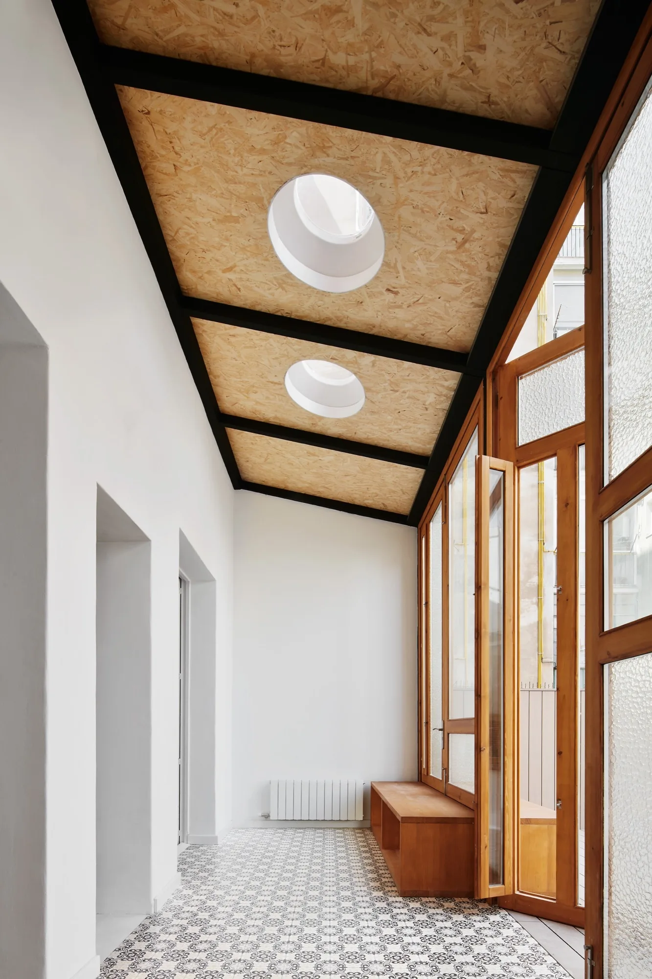 A transition corridor with a sloped OSB ceiling, circular skylights, a wood-framed window wall, and geometric tiled flooring.