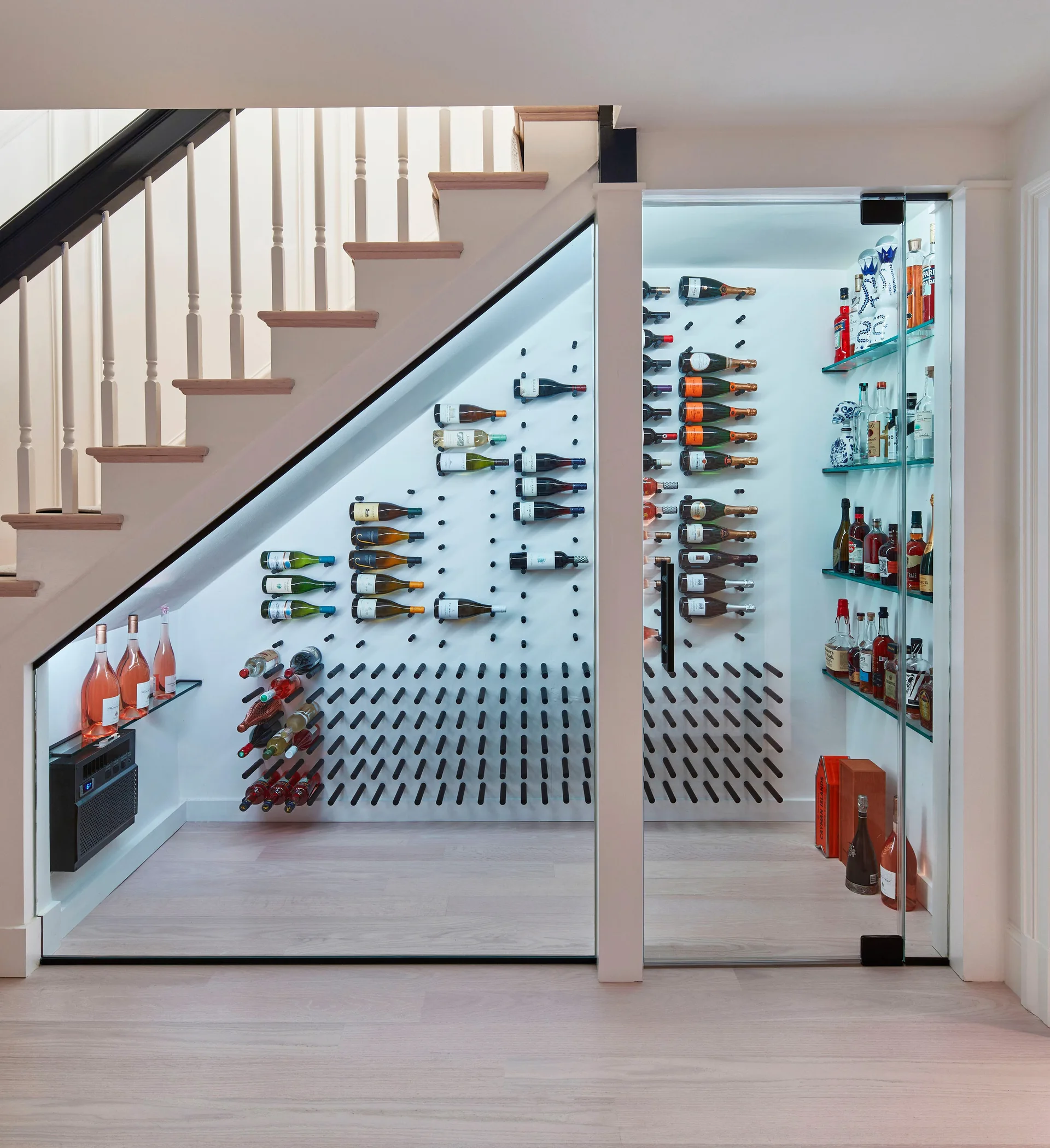 A glass-enclosed wine cellar built under a wooden staircase features a white wall with black peg racks, liquor shelves, and bright LED lighting.