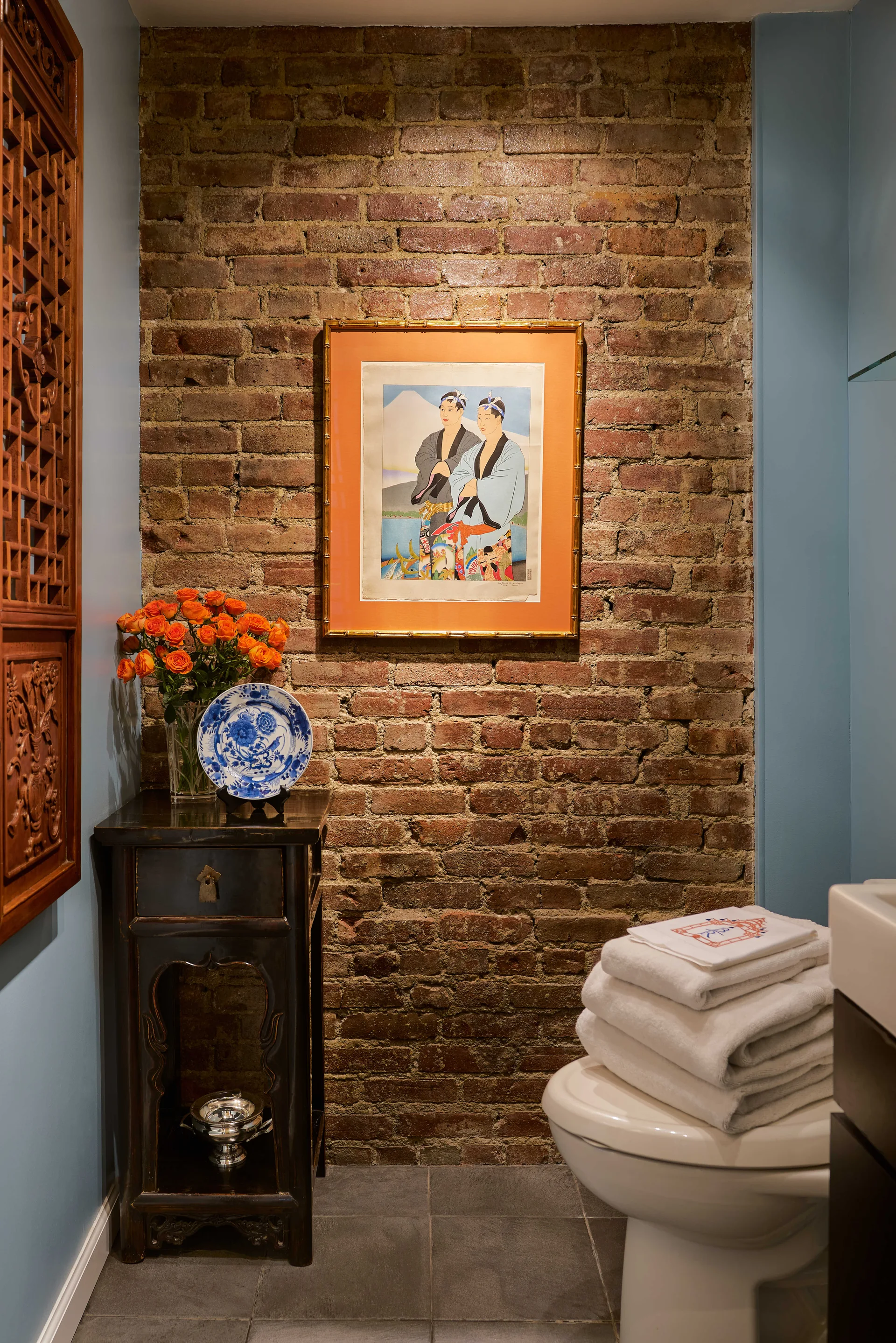 A powder room features a textured red brick wall with framed art, adjacent to blue walls, a dark wood side table, and a white toilet.