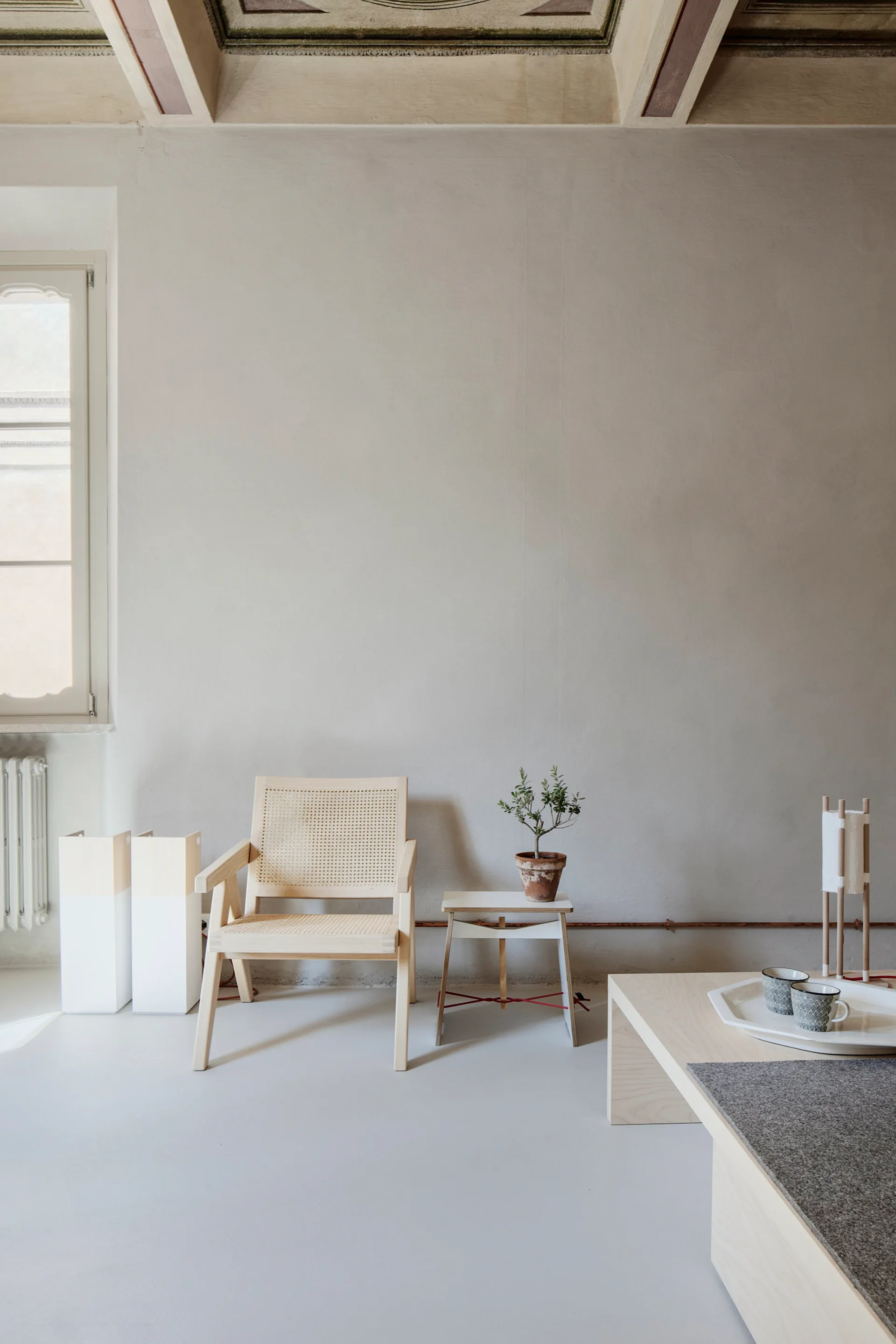 A light wood cane armchair sits next to a small side table in a minimalist living room with a low coffee table and decorative ceiling beams.