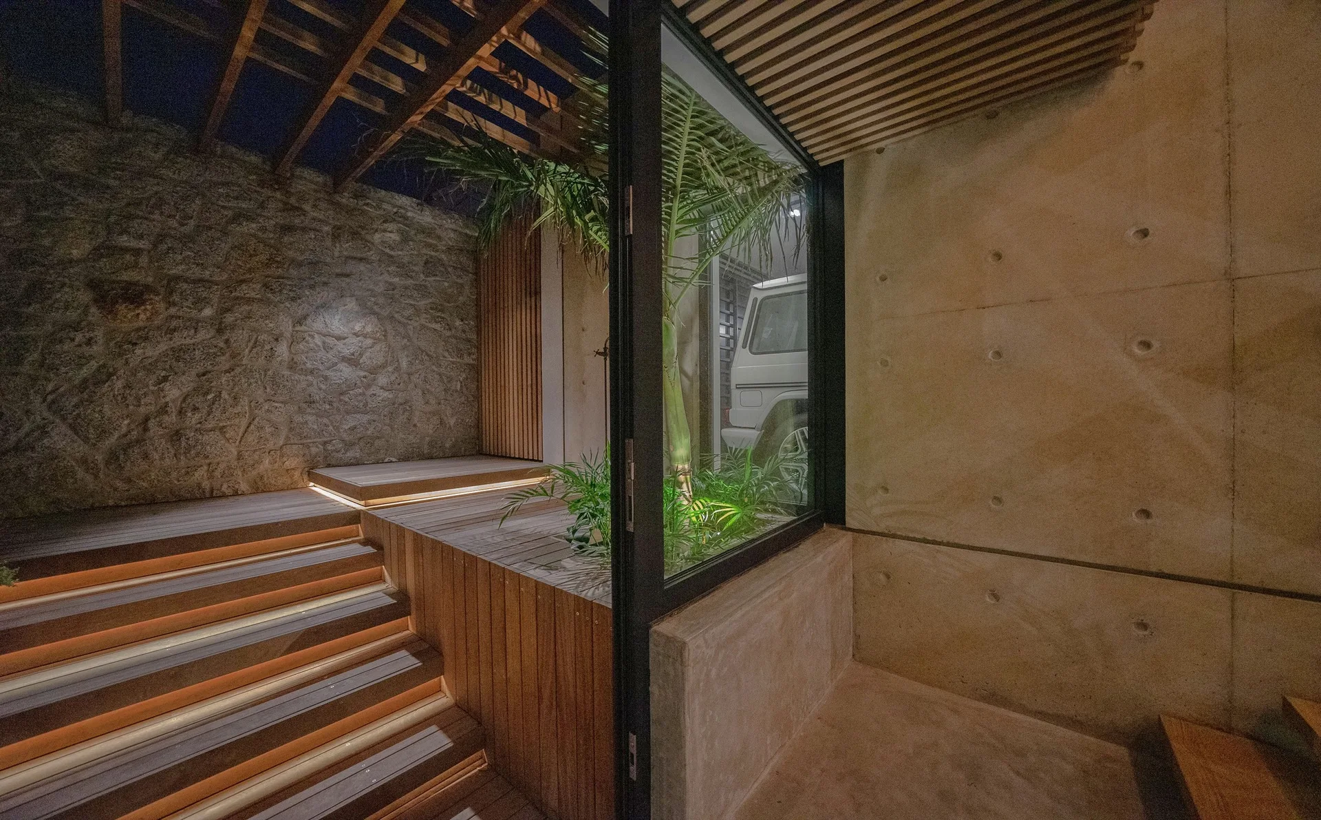 An outdoor patio featuring wooden stairs with integrated warm lighting, a rustic stone wall, and a smooth concrete wall under a slatted ceiling.