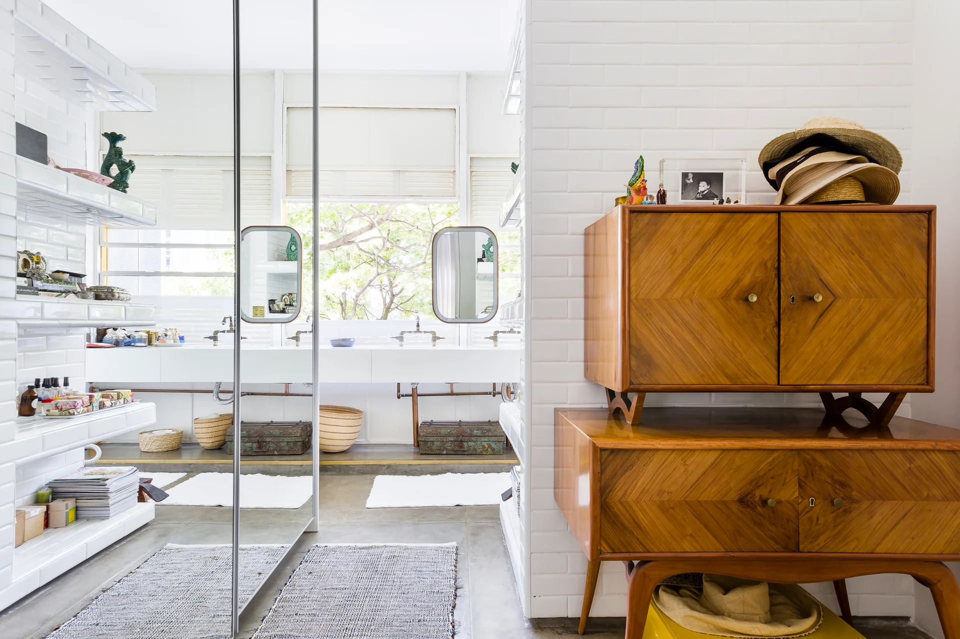 A bright bathroom with white glazed brick walls, a mid-century wood cabinet, and a double vanity reflected in a large mirror panel.