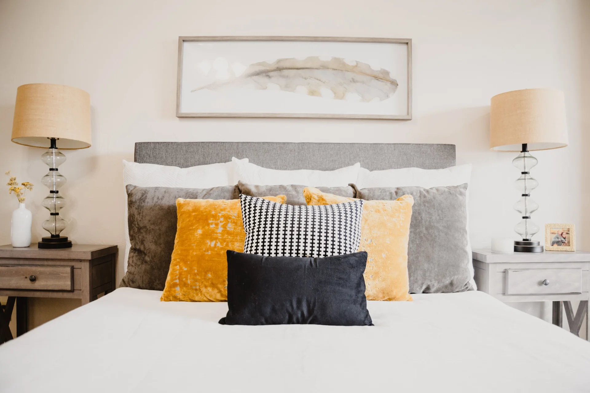 A grey upholstered headboard is flanked by matching nightstands with glass table lamps and decorated with mustard yellow, black, and grey pillows.