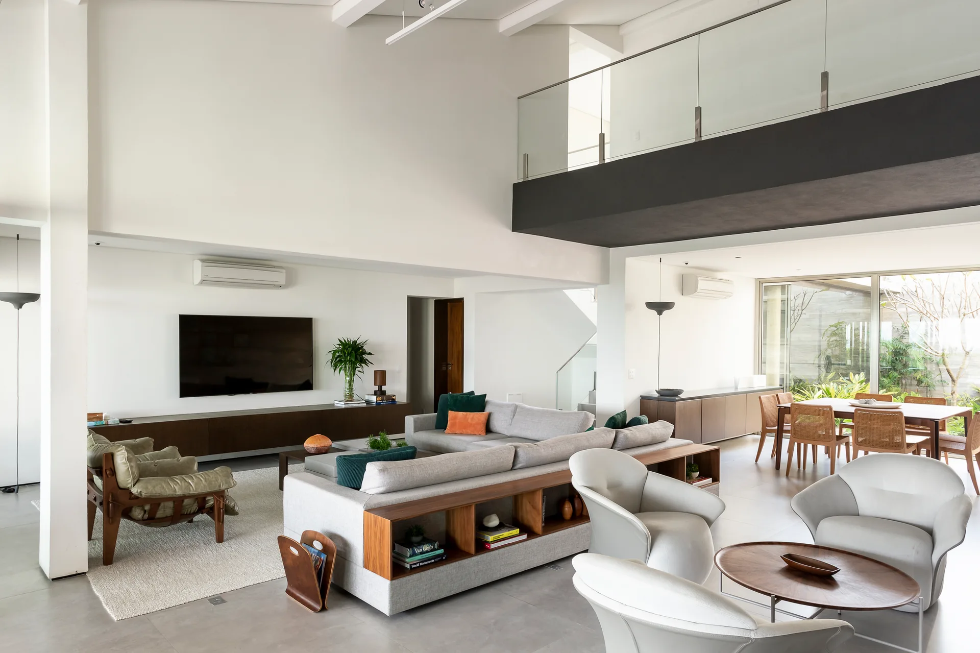 A double-height living room features a grey sectional sofa with built-in wood shelving, a glass mezzanine, and a dining area.