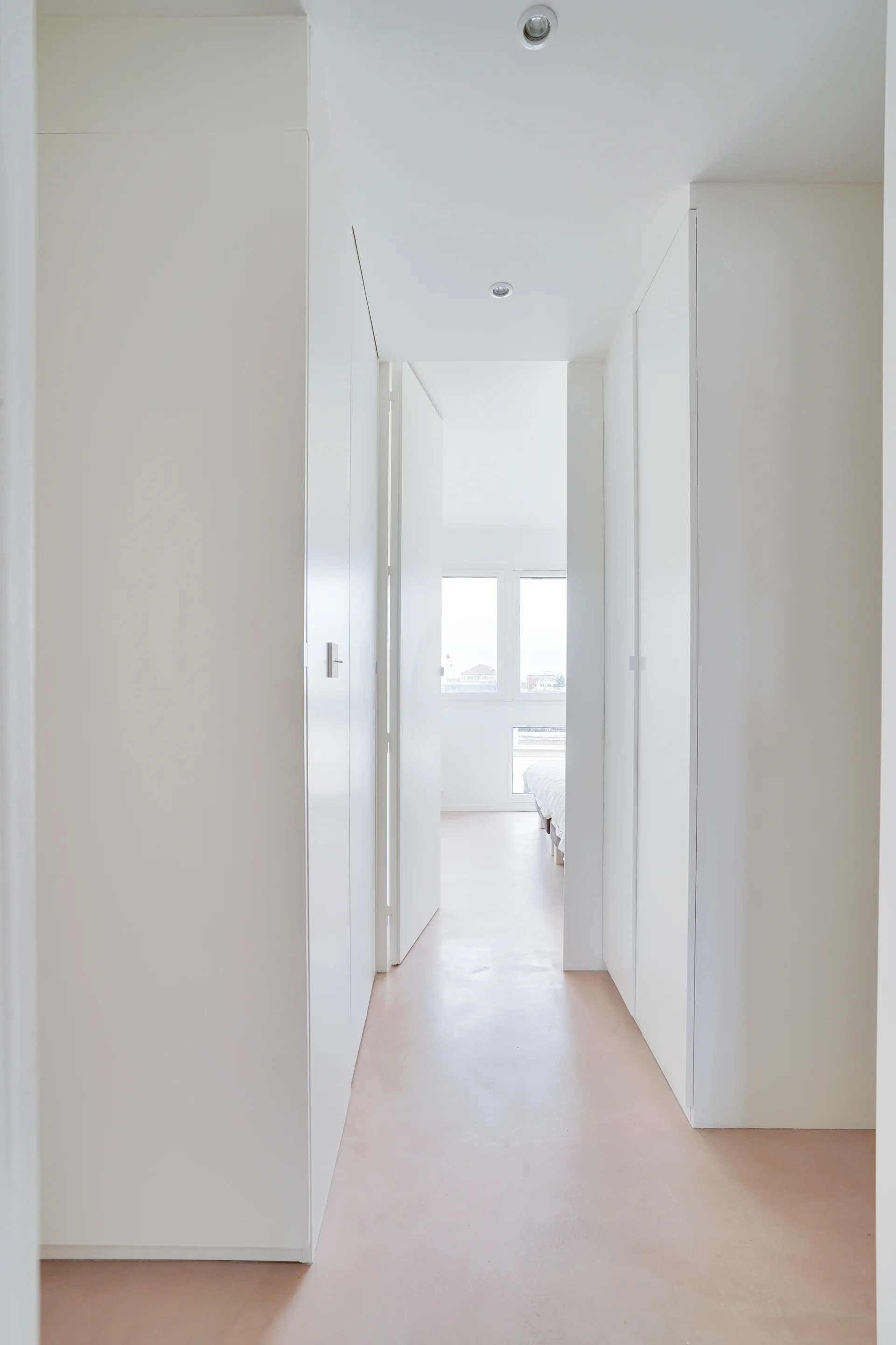 A minimalist white hallway features floor-to-ceiling built-in storage closets, recessed ceiling lights, and a pale pink floor leading into a bedroom.