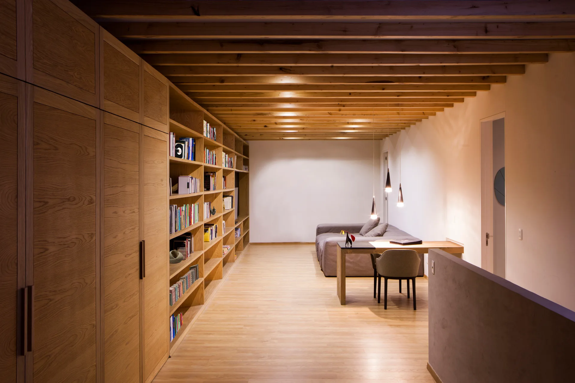 A modern long room with a large oak built-in bookshelf, exposed wooden ceiling joists, and a desk under three hanging pendant lights.
