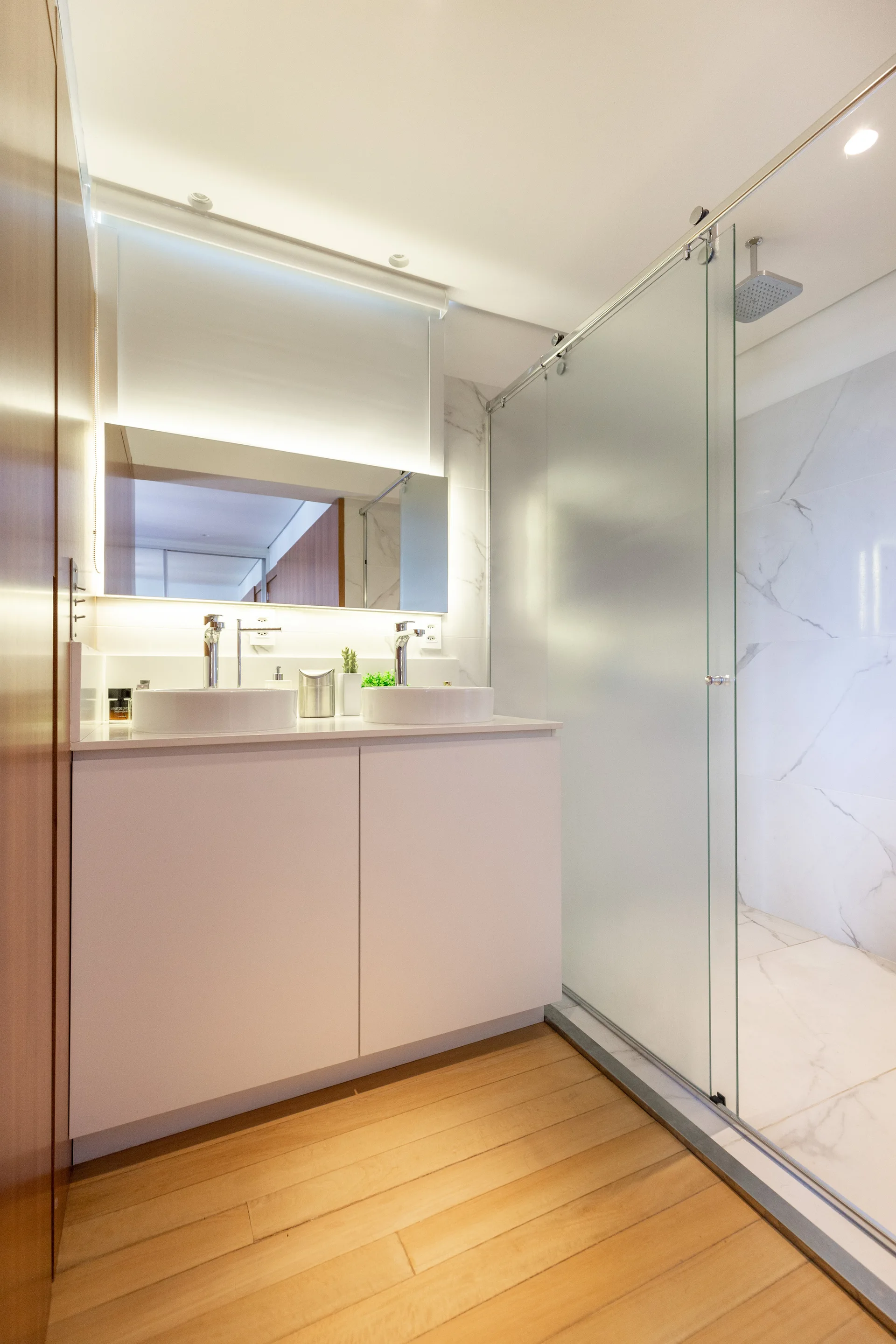 A modern bathroom features a white double vanity with vessel sinks, a backlit mirror, wood flooring, and a frosted glass shower enclosure.