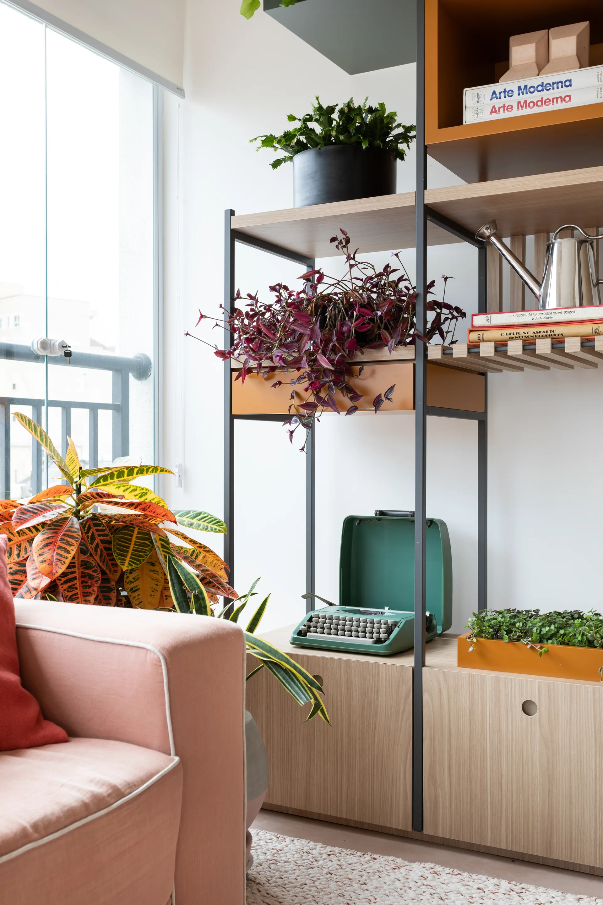 A tall wooden and black metal shelving unit styled with leafy green plants stands beside a light pink sofa.